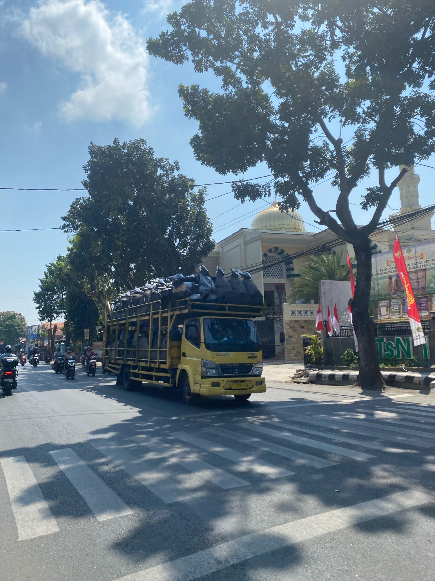 This is a vertical, medium-shot, eye-level, outdoor photograph captured on a sunny day in Kediri City, Indonesia. The scene depicts a street with a pedestrian crossing in the foreground, marked by white stripes on asphalt. A yellow truck, laden with many dark, tarp-covered bundles, is moving from left to right across the frame, partially obscuring the view of the street and buildings behind it. The truck has a cage-like structure supporting the cargo and appears to have people inside the truck bed.
In the middle ground, several motorcycles are seen traveling in the same direction as the truck. To the right of the truck, a building with a golden dome and minaret, likely a mosque, is visible. Red and white Indonesian flags are displayed near the mosque. A large tree with dense foliage stands between the truck and the mosque, casting shadows on the road. The background features more trees and buildings, suggesting an urban or suburban environment. The sky is blue with scattered white clouds. The overall impression is a busy street scene in a city.