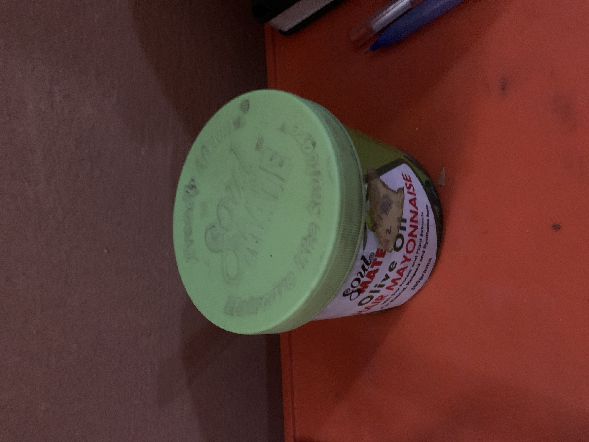 A green plastic tub of Soul Mate Olive Oil Hair Mayonnaise sits on a bright orange surface. The tub is mostly visible, with its label showing the product name and ingredients. The lid of the tub has a raised, embossed pattern of text. The background is a textured, reddish-brown wall. Two pens, one blue and one red, are positioned to the right of the tub, partially obscuring a black notebook.