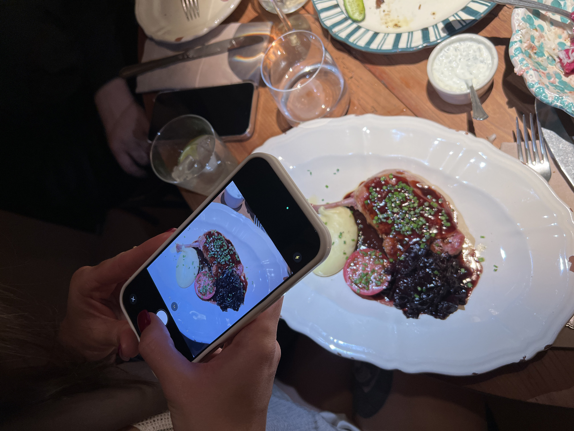 A person is holding a smartphone to take a picture of a plate of food on a wooden table. The plate contains what appears to be meat with a dark sauce, mashed potatoes, and a sliced vegetable. Other plates and glasses are on the table, some partially eaten. The environment suggests a restaurant setting. There is no indication this image was taken in Moscow, Russia.