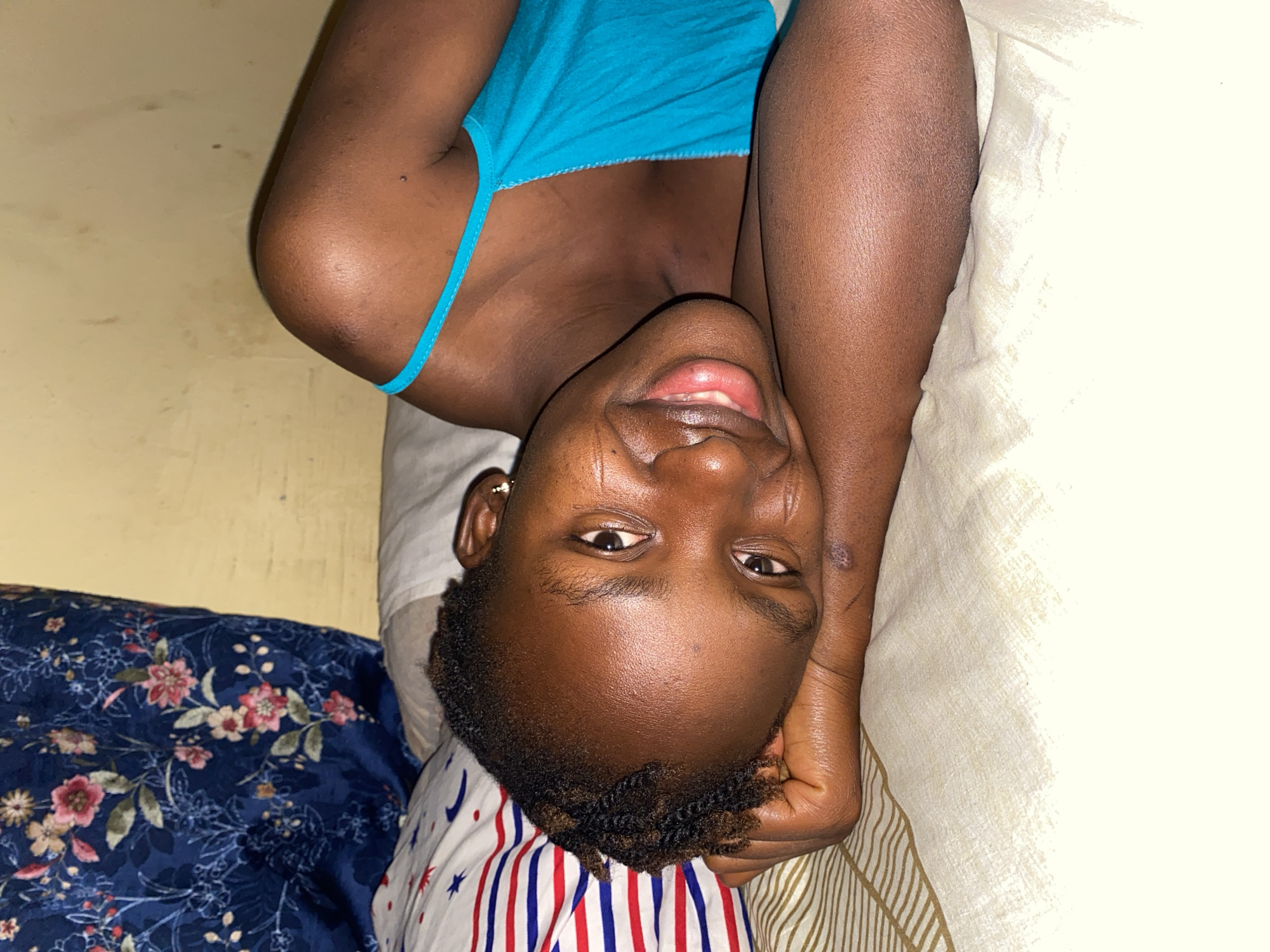A woman with dark skin lies on a bed with her head resting on pillows with different patterns. She is wearing a blue tank top. The scene appears to be indoors, possibly in a residential area in Kuru Dungus, Nigeria. The wall behind her is a light yellowish color. The lighting is dim. The woman is smiling.