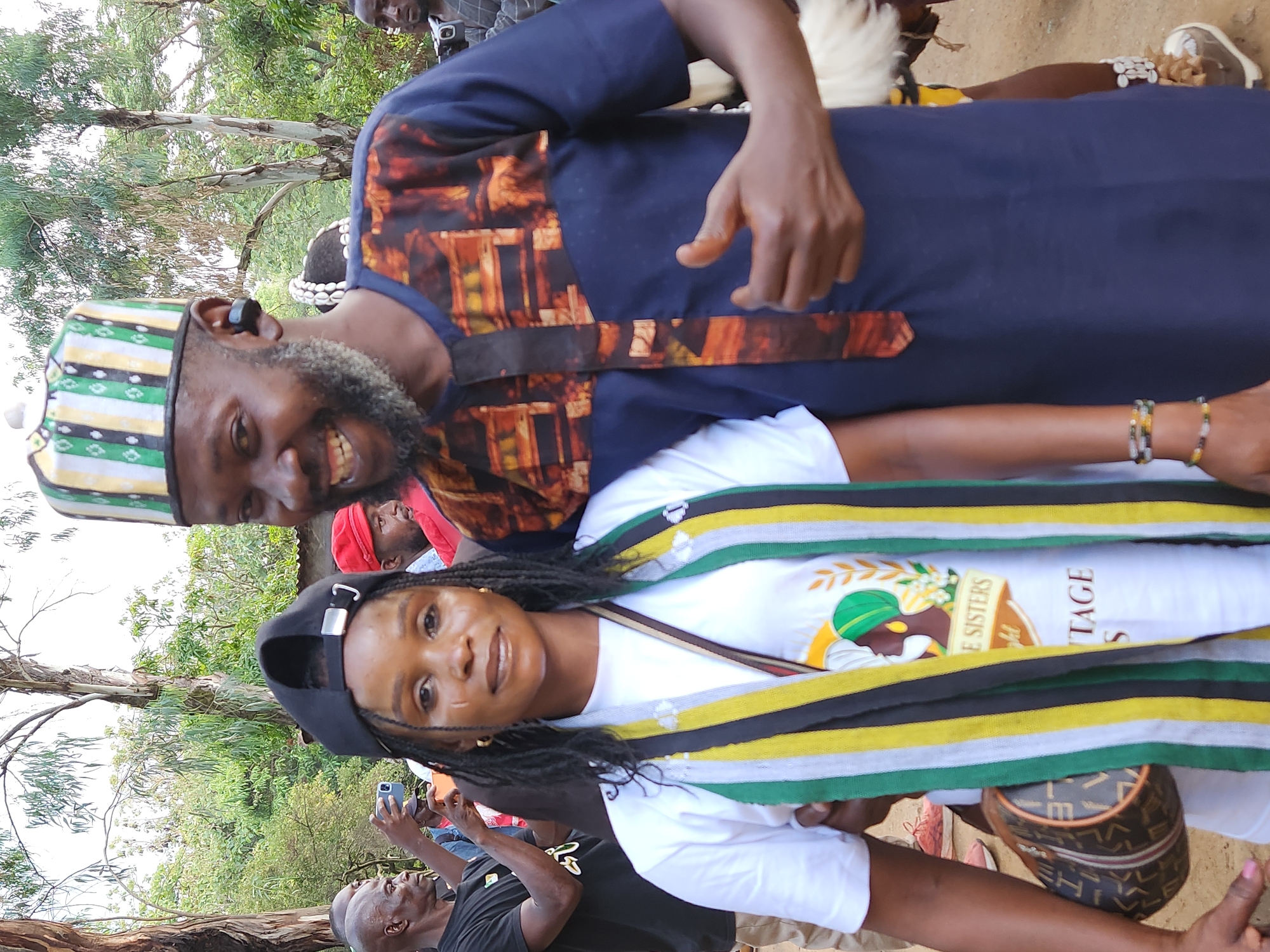 This is a medium shot photograph of a group of people outdoors, likely in Jos, Nigeria, given the context of the image. In the foreground, two individuals are prominently featured. On the left is a woman wearing a white t-shirt, a black baseball cap, and a scarf with green, yellow, and black stripes. She has braids and is wearing gold hoop earrings. A small, patterned handbag is slung across her body. On the right is a man with a beard, wearing a navy blue garment with an orange and brown patterned fabric accent. He has a distinctive hat with green, white, and yellow stripes. Both individuals are smiling and looking towards the camera.
Behind them, several other people are visible, some holding cameras or phones, suggesting an event or gathering. The background consists of trees and foliage, indicating an outdoor setting. The ground appears to be dirt. The overall atmosphere seems festive and communal.