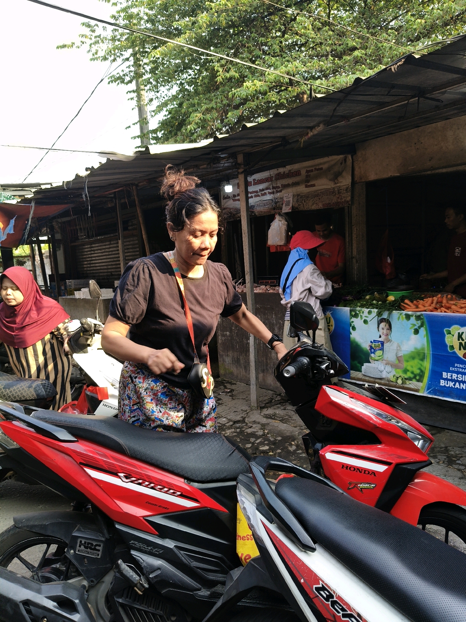 In Setiamekar, Indonesia, a woman in a black shirt and colorful patterned skirt stands next to a red Honda motorcycle, appearing to adjust something on the handlebars. To her left, another woman wearing a maroon hijab and a striped dress is seated on a motorcycle. Behind them, a market stall is visible with various produce, including carrots and green vegetables, laid out on a counter. A banner featuring a woman holding a cleaning product and text in Indonesian is displayed at the stall. Several other people are present in the background, some appearing to be vendors and customers. The scene is outdoors, under a tin-roofed structure, with trees visible in the background.