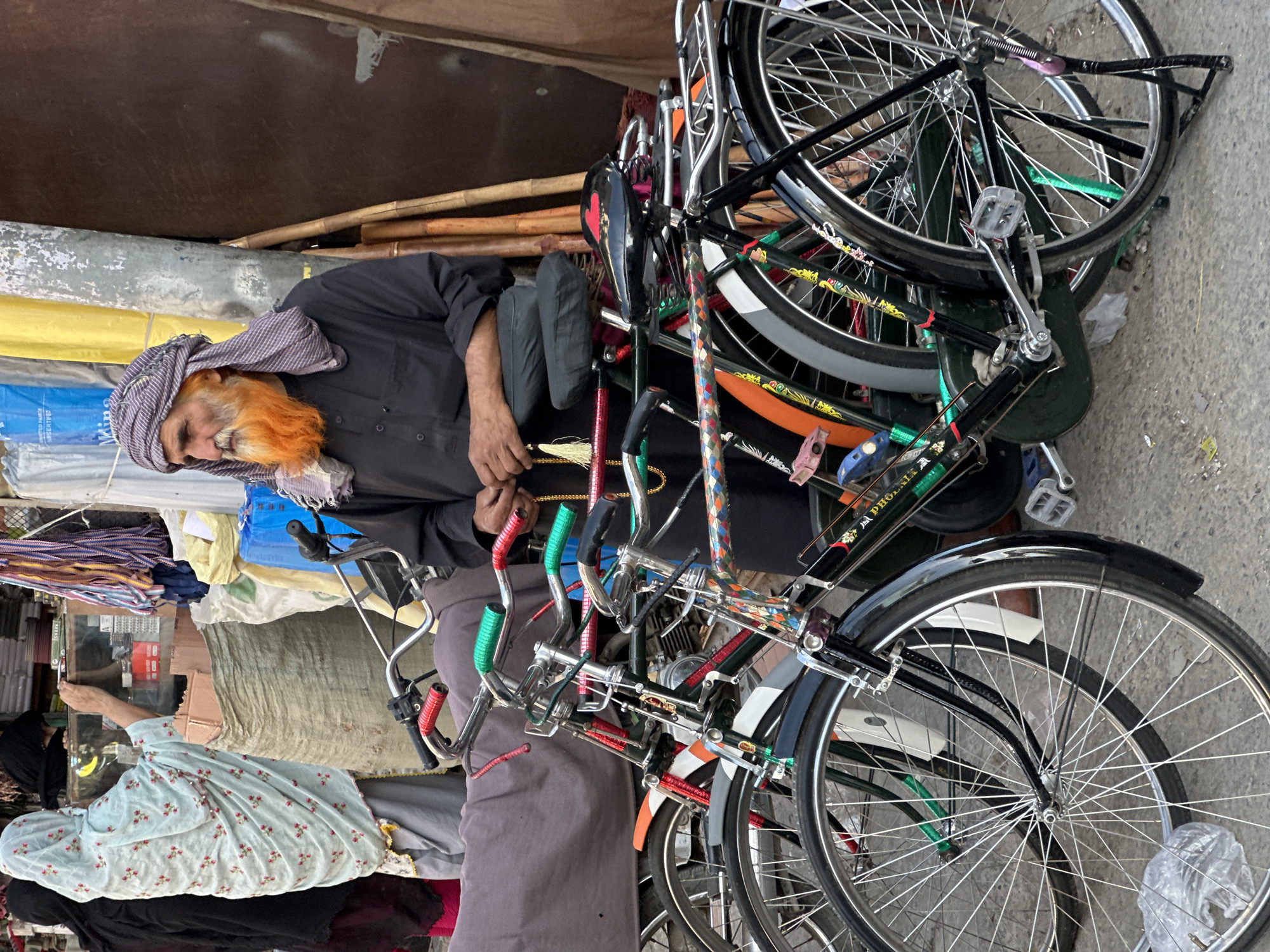 In Quetta City Tehsil, Pakistan, an elderly man with a vibrant orange beard and a purple and white checkered turban stands beside several bicycles. He wears a black long-sleeved shirt. His hands are clasped in front of him, and he holds a string of prayer beads. The bicycles, parked in a row, have various colored handlebar grips, including red and green, and some have decorative patterns on their frames. In the background, a woman in a light blue patterned headscarf and a grey skirt is visible, seemingly interacting with a stall or shop. The environment appears to be a bustling market or street scene, with assorted goods and materials piled up.