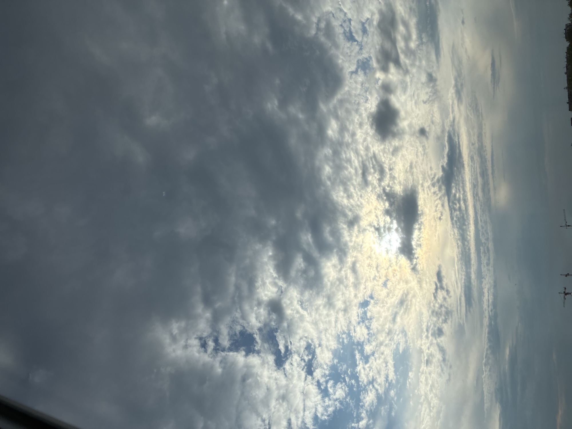 An outdoor, full-frame, eye-level shot captures a cloudy sky above a cityscape. The sky is filled with a mix of dark and light clouds, with patches of sunlight breaking through, creating a dramatic and somewhat ominous atmosphere. The clouds are varied in texture and density, with some appearing wispy and others more solid and textured. The lower portion of the image reveals the silhouettes of buildings and construction cranes against the lighter horizon, suggesting an urban or industrial environment. The lighting is subdued, with the sun's rays diffused by the cloud cover, casting a soft glow in some areas and deep shadows in others. The overall impression is one of a dynamic sky in an urban setting, possibly during late afternoon or early morning.