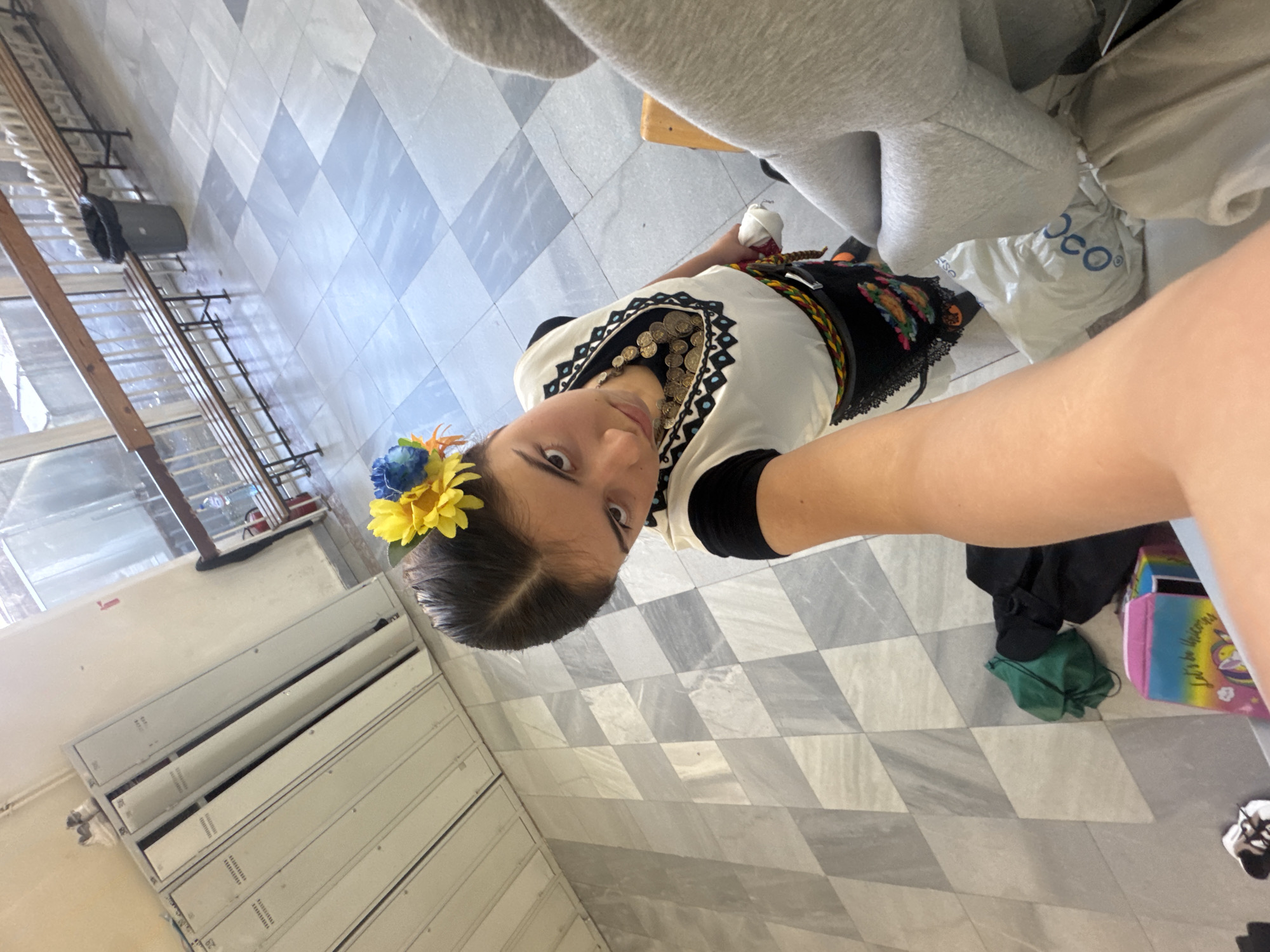 A young woman is depicted in a selfie-style photograph, seemingly indoors, possibly in a school environment given the lockers visible along one wall. She is wearing a traditional embroidered outfit, accented with a floral headpiece. The environment includes a checkered tiled floor, lockers, and miscellaneous items such as bags and clothing scattered around her feet. The setting suggests an informal, candid moment captured within an institutional building.