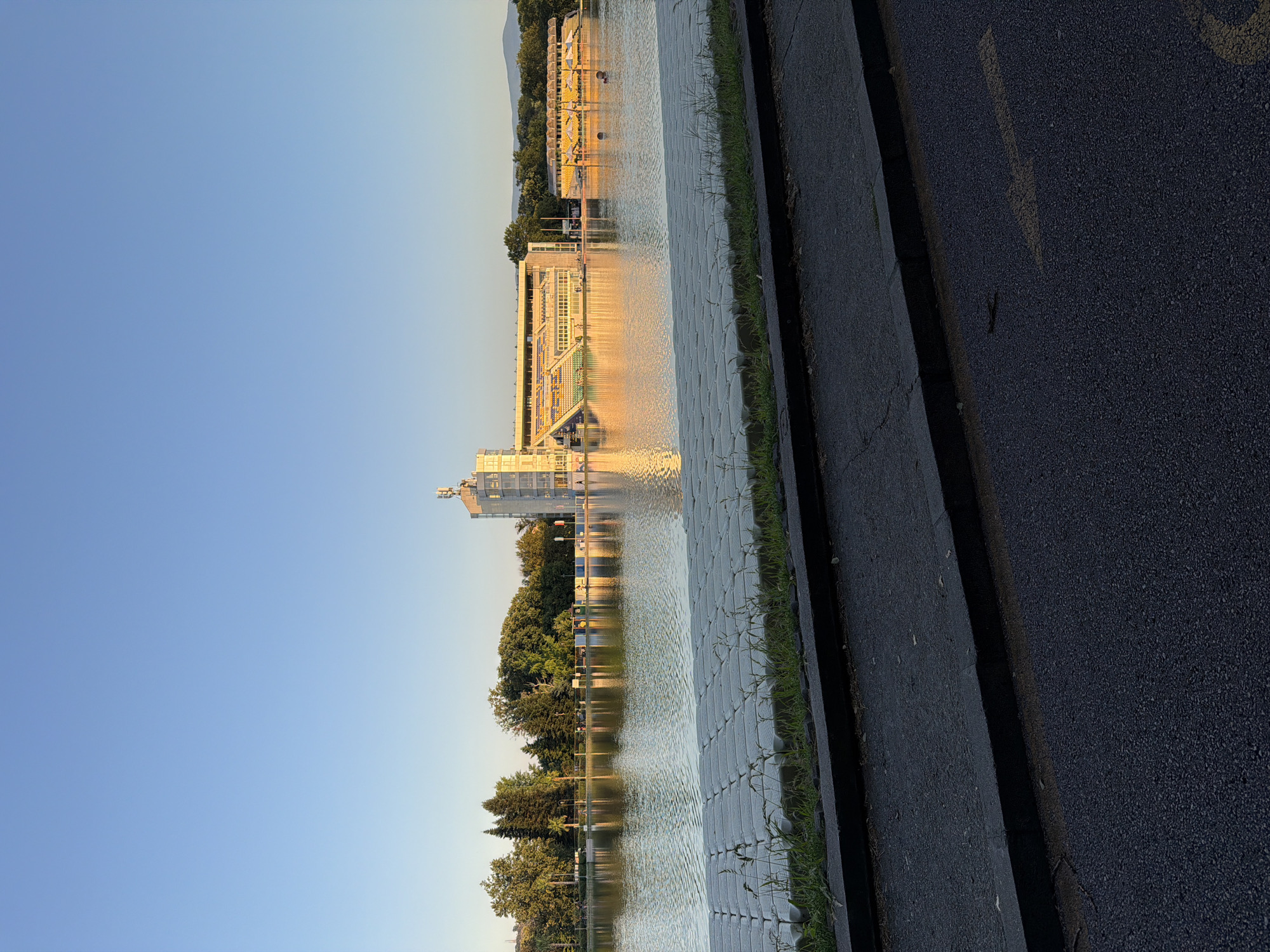 This image depicts the Plovdiv Rowing Canal in Plovdiv, Bulgaria, at sunset. The sun's golden light reflects off the calm water, creating a serene atmosphere. On the far bank, a large, modern building with tiered seating, likely a stadium or sports facility, is visible. The building is illuminated by the setting sun, highlighting its architectural features. The canal is lined with trees and some smaller structures with yellow awnings. The foreground shows a paved walkway or road with a grass border. The sky is clear and blue, indicative of fair weather. There are a few individuals visible on the far bank and in the water, suggesting the location is accessible and utilized by the public. The overall scene is tranquil and picturesque.