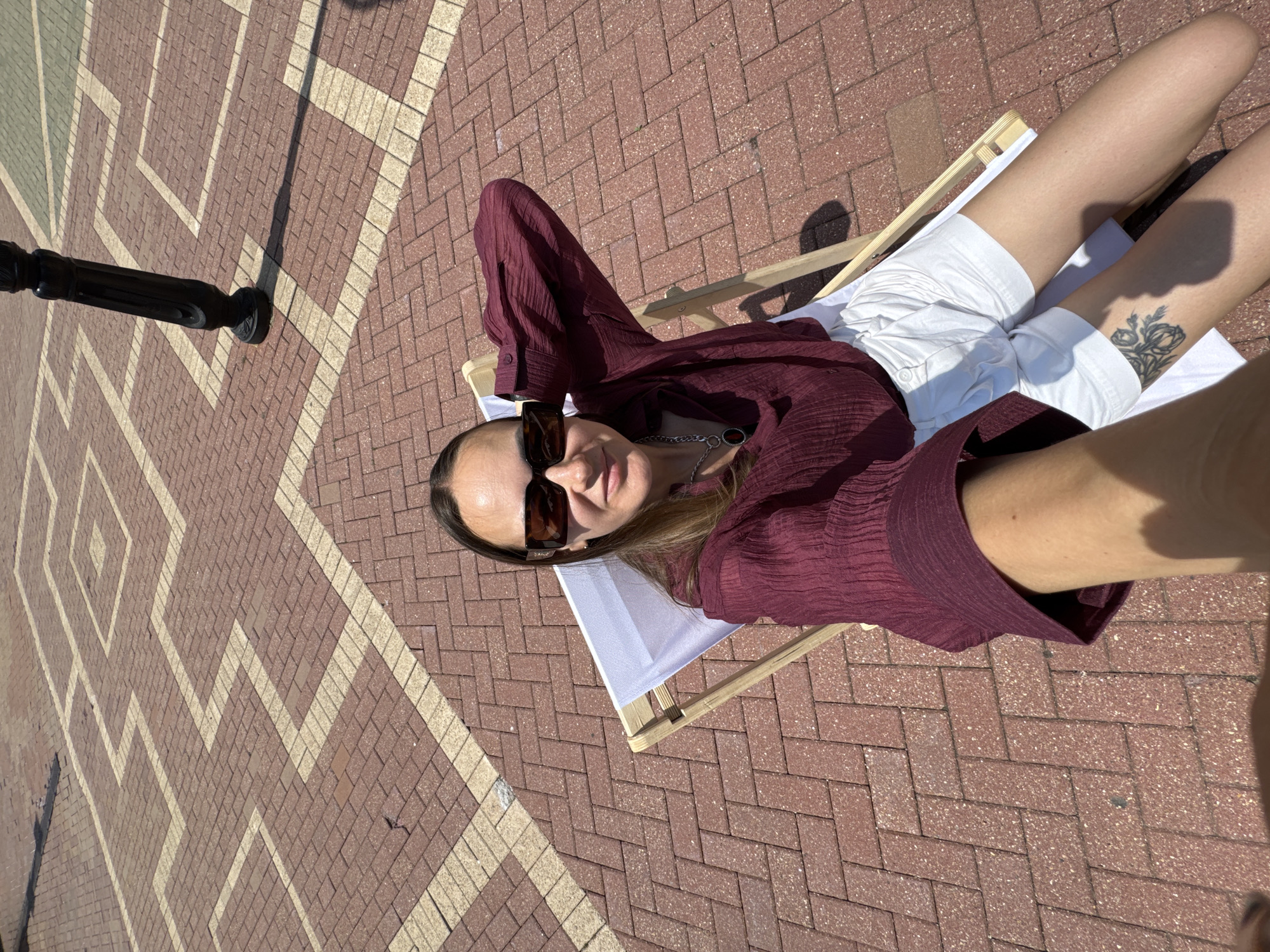 A woman is lying on a deck chair on a brick-paved area. She is wearing a maroon blouse and white shorts. She is also wearing sunglasses. The sun is shining, and there is a shadow of the woman on the ground. The woman is looking at the camera and smiling. The background is a brick-paved area with a geometric pattern. There is a black pole on the left side of the image, which might be a lamppost. The woman has a tattoo on her left thigh. The time of day appears to be daytime, likely sunny weather, given the shadows. The setting appears to be an outdoor public space, possibly a park or a plaza, given the paving and lamppost. The woman seems relaxed and happy, enjoying the sunny weather. There is no visible text in the image.