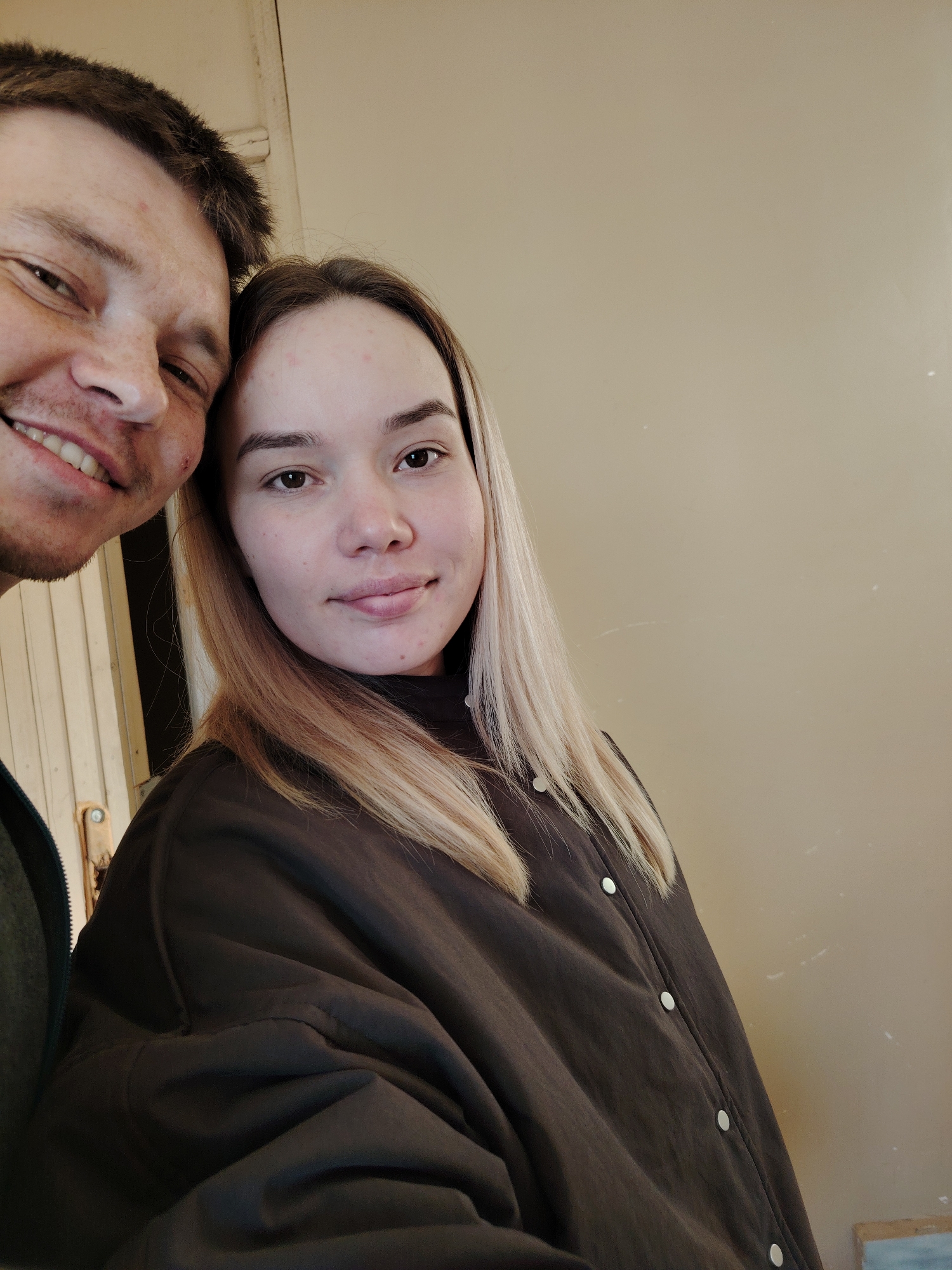 A close-up shot captures two people, a man and a woman, inside a building with a beige wall. The woman has light brown hair with blonde highlights and wears a dark brown jacket with visible white buttons. The man, positioned on the left side of the frame, has short brown hair and is smiling. The location appears to be inside, potentially Марковский, Russia.