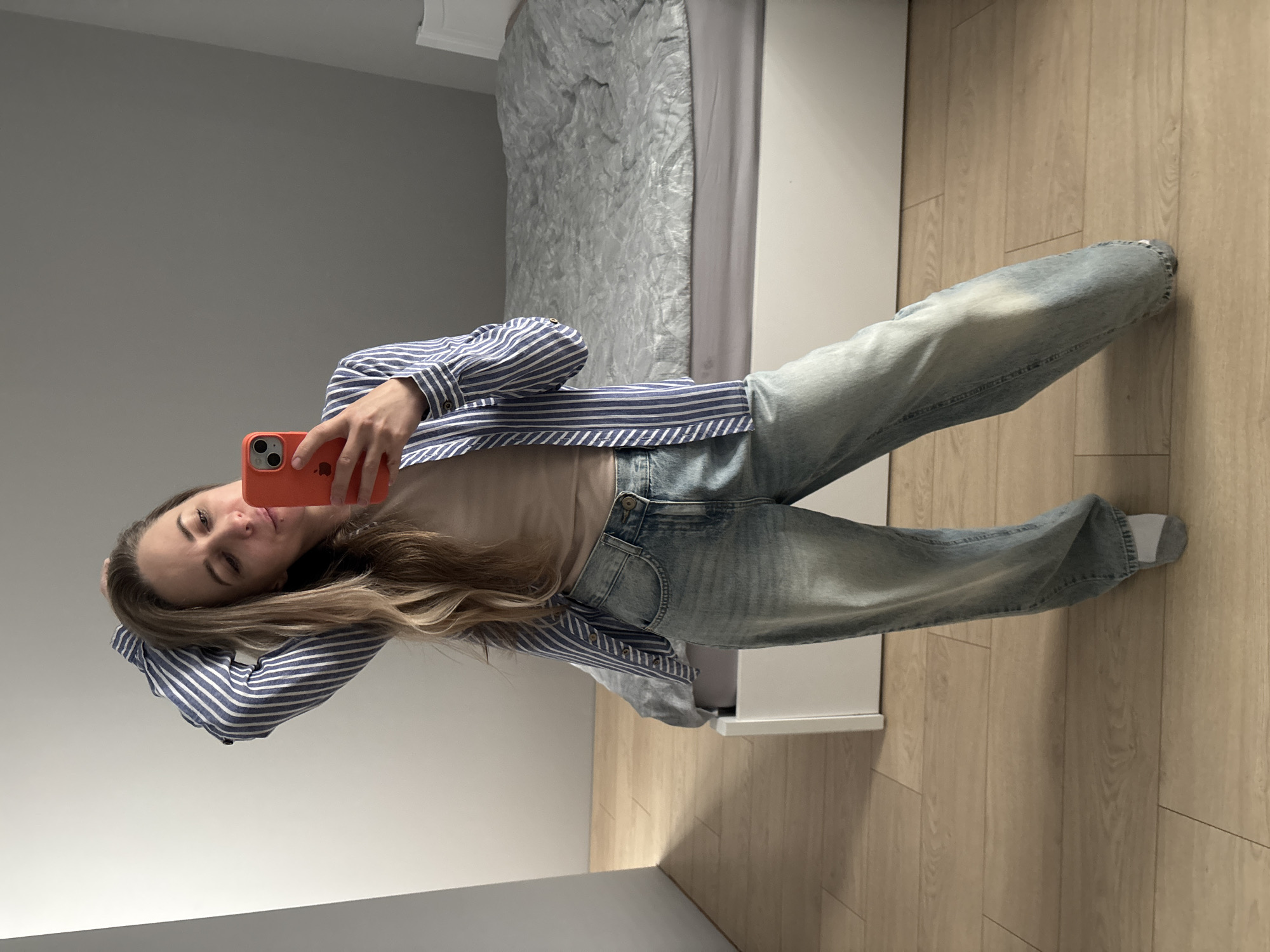 A woman stands indoors in front of a bed with a grey comforter. She is taking a selfie using an orange phone. She wears jeans, a tan shirt, and a blue and white striped long-sleeved shirt. The room has light-colored wooden flooring and grey walls. The exact location in Chaikovsky, Russia, is not discernible from the image.
