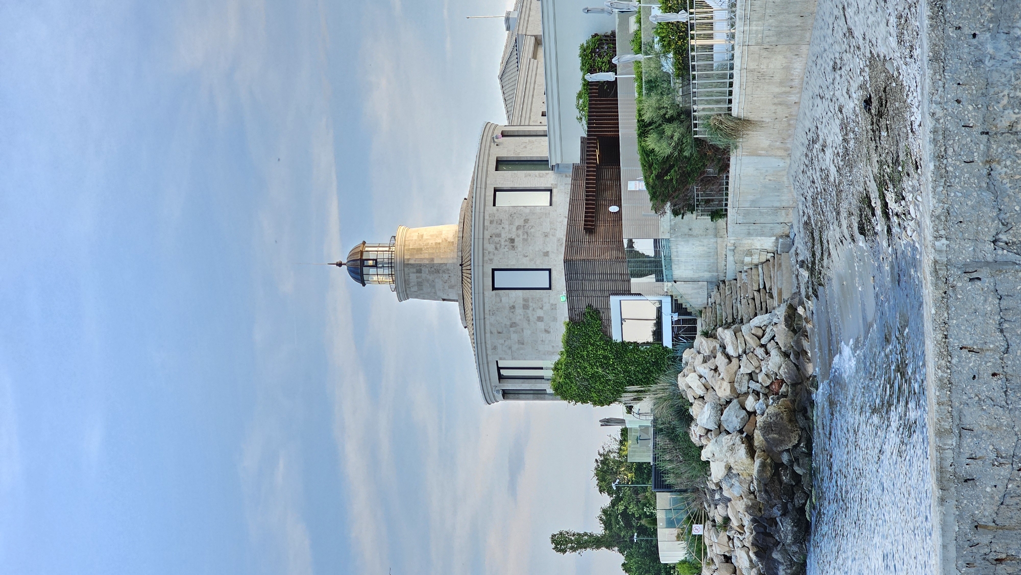 The image shows a lighthouse located in Varna, Bulgaria. The lighthouse is a tall, cylindrical structure with a glass dome at the top. It is made of stone and has several windows. The lighthouse is located next to a building with ivy and what appears to be a beach, with water and rocks on the right side of the image. The sky is blue with white clouds.