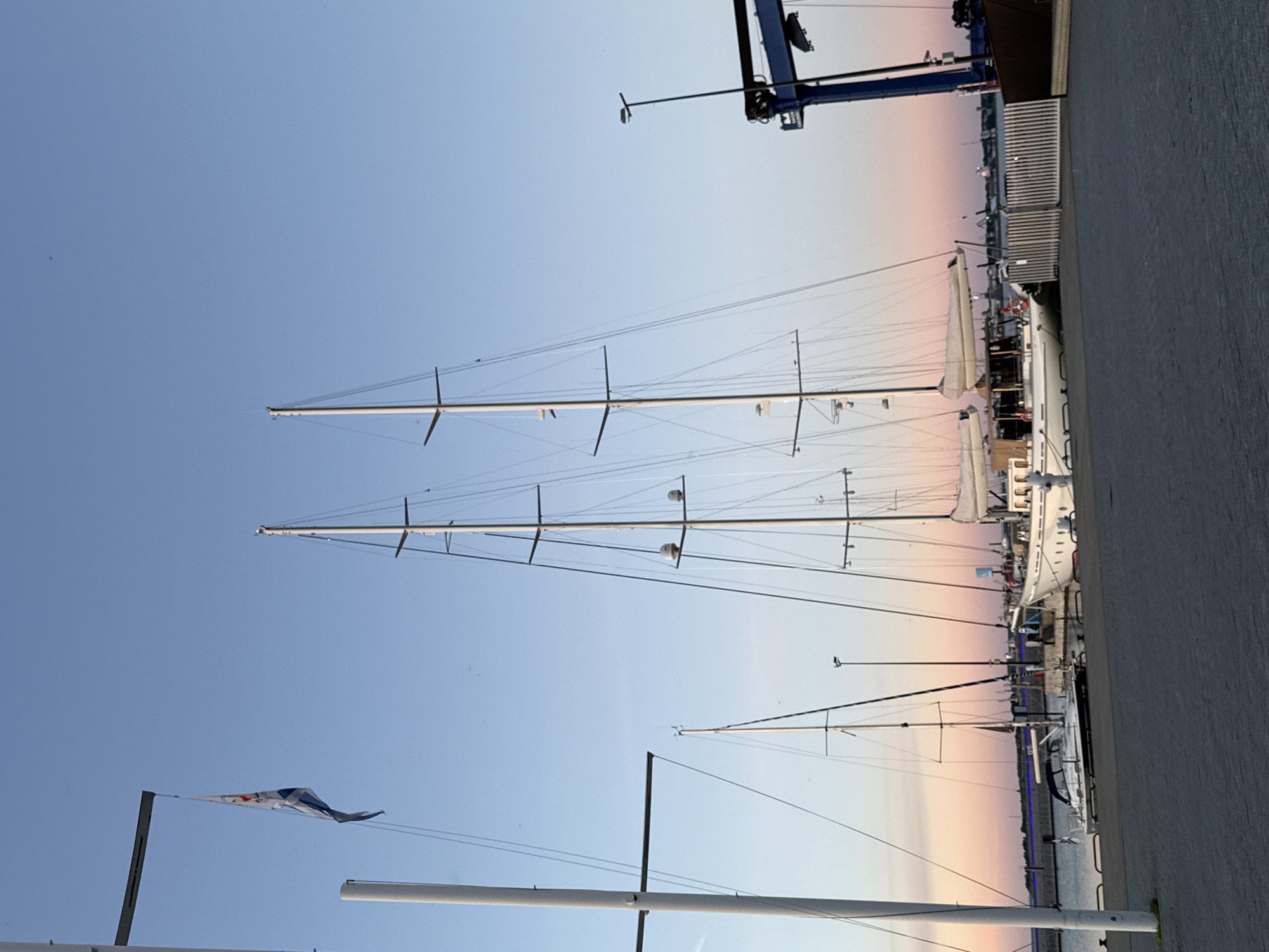The image shows a harbor scene in Tallinn, Estonia, at dusk. The sky is a gradient of blue to pink, indicating the setting sun. Several sailboats are docked along a paved walkway. The largest sailboat in the foreground has white sails and a wooden deck. To its left, a smaller, sleeker sailboat with a dark blue canopy is moored. In the background, a large blue crane is visible, suggesting a shipyard or industrial area. A flag with a blue and white pattern flies from a flagpole on the left side of the image. The overall atmosphere is serene and tranquil.