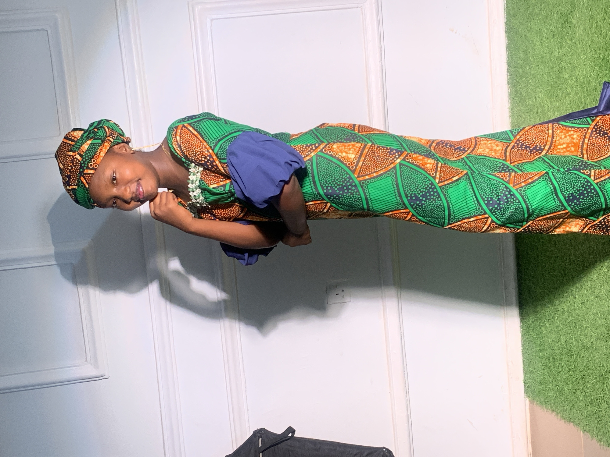 The photograph depicts a young girl in Barakin Akawo, Nigeria. She is wearing a traditional African dress with a green and orange patterned fabric, complemented by blue sleeves and a matching headwrap. A white beaded necklace adorns her neck. The girl is posing with a slight smile, her hand near her chin. The background consists of a white paneled door and a section of artificial green turf. In the lower part of the frame, there is a partially visible black object, possibly a piece of furniture or equipment. An electrical outlet is also visible on the white door.