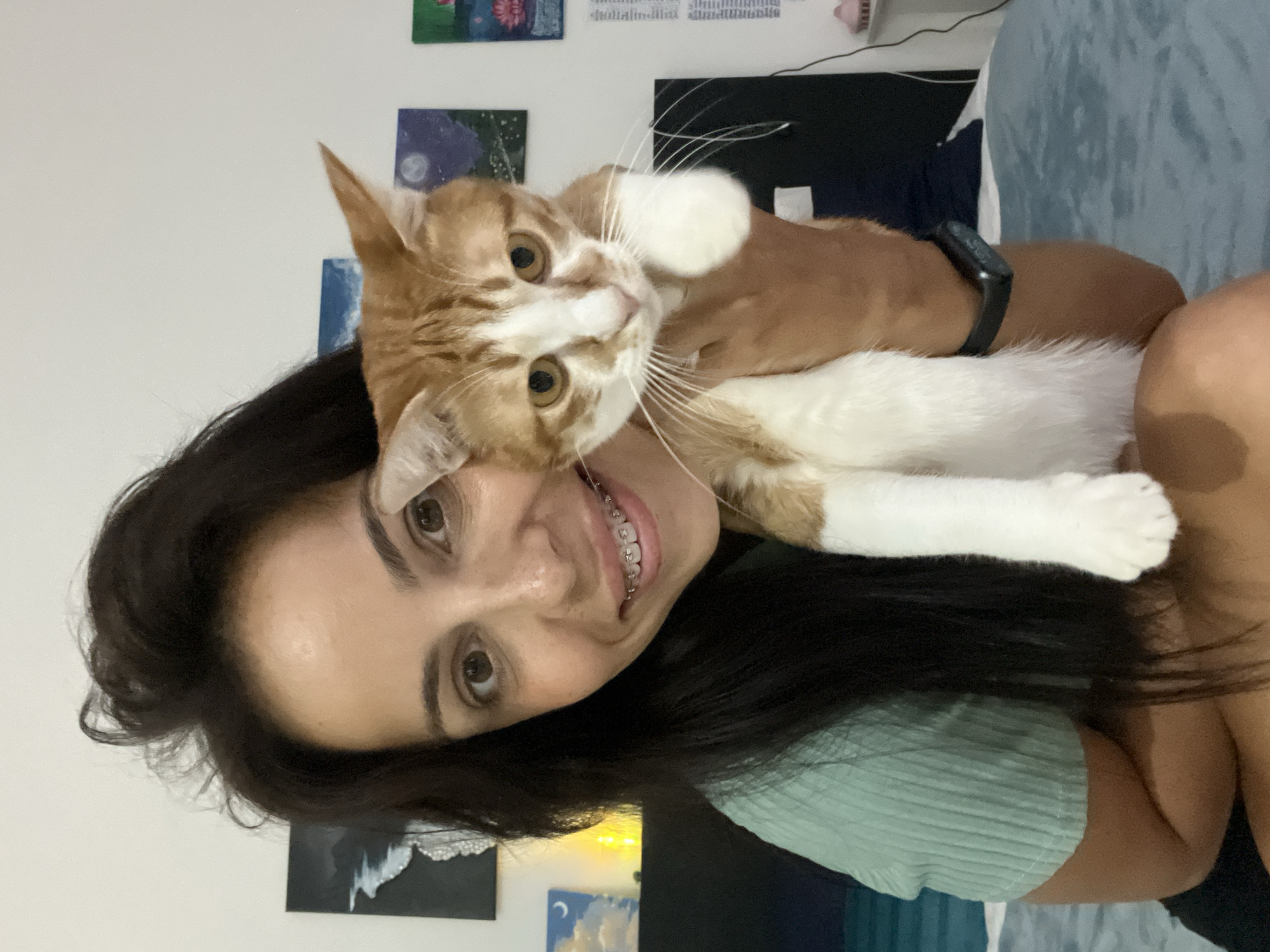 An intimate, rotated portrait captures a young woman and her orange and white cat indoors in Kuta Selatan, Indonesia. The woman, with long dark hair, smiles broadly at the camera, revealing braces on her teeth. She wears a light green ribbed top and a black smartwatch on her left wrist, which partially displays "WED 15" and other illegible text.
Perched on her shoulder or chest, the cat looks directly at the camera with wide, golden eyes and prominent white whiskers, its white paws and chest fur contrasting with its orange patches. The woman's joyful expression and the cat's direct gaze convey a warm, affectionate interaction.
The background shows a plain white wall decorated with several small artworks, including paintings of a night sky with a crescent moon and stars, and what appears to be a floral design. Fairy lights are visible in the lower background, contributing to a cozy ambiance. A dark object, possibly a speaker or shelf, is also partially visible, along with a light blue fabric item in the upper right. The scene suggests a casual, personal moment captured under indoor lighting.