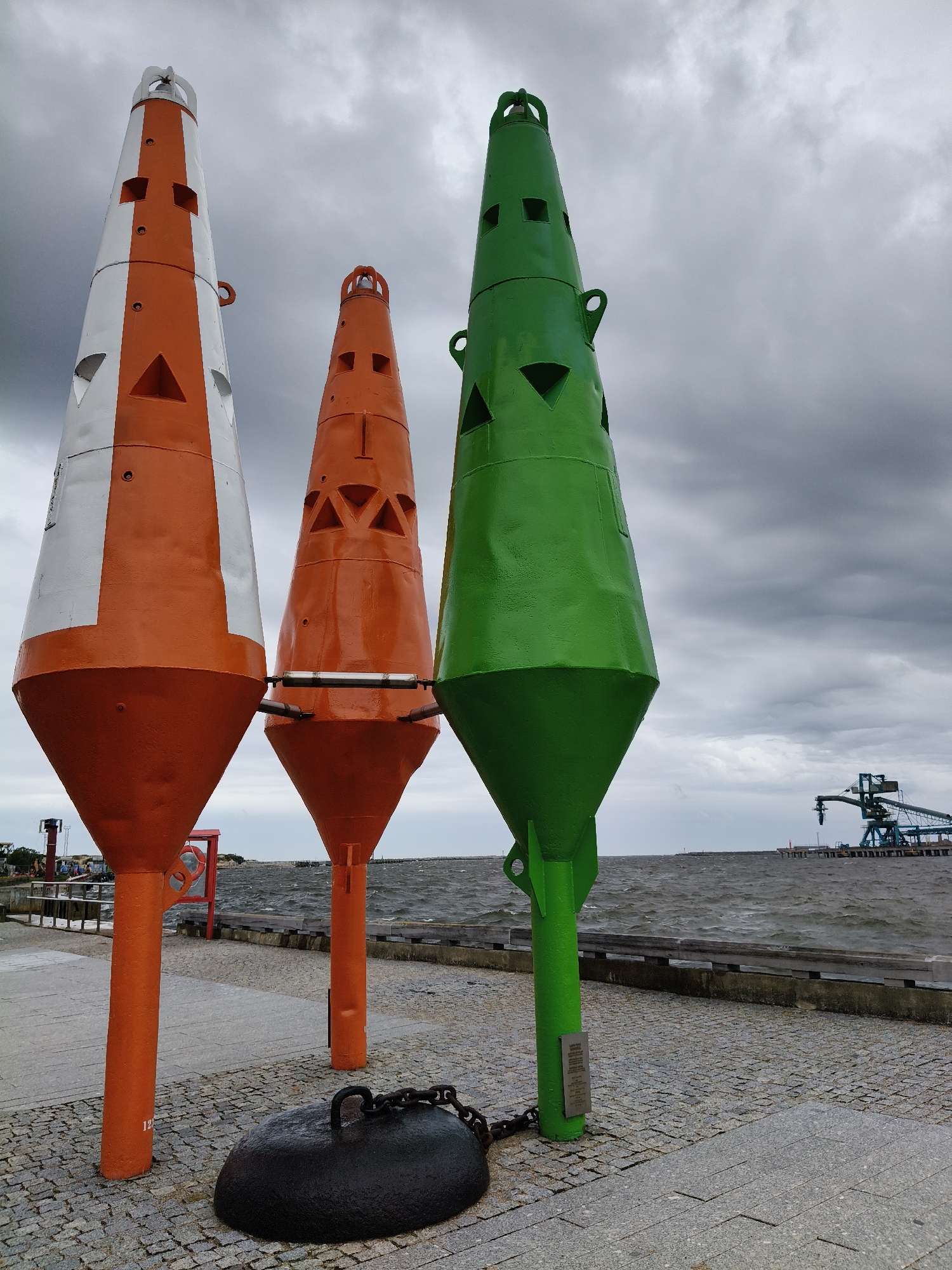 Three large, conical buoys stand in a row on a paved walkway by the sea. The buoy on the left is orange and white, the middle buoy is orange, and the buoy on the right is green. Each buoy has an open top and several square or triangular openings along its sides. A large, dark, anchor-like object with a chain attached rests on the ground in front of the buoys. In the background, the choppy, grey sea stretches to the horizon under a cloudy sky. A pier with industrial structures, including a conveyor belt system, is visible in the distance. The scene appears to be located at the Ventspils Port in Ventspils, Latvia.