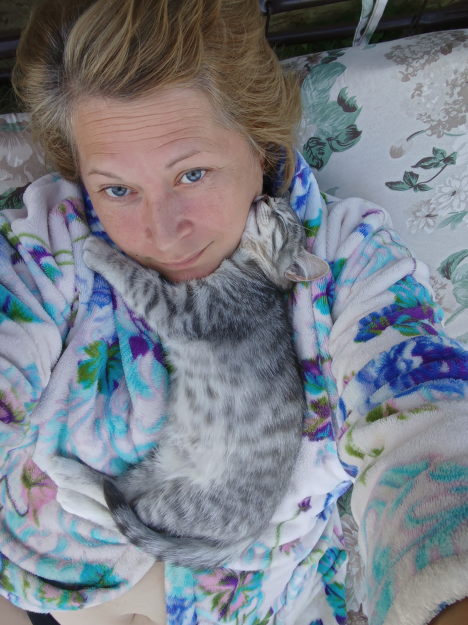 A close-up overhead shot shows a woman with blonde hair and blue eyes lying on a floral patterned cushion, wearing a fluffy robe with a floral print. A small, grey tabby kitten is nestled on her chest, with its head resting on her chin. The background is blurred, but appears to be outdoors. The image is taken from above, looking down at the woman and kitten. There is no discernible indication of the location in Ivanovskoe, Russia.