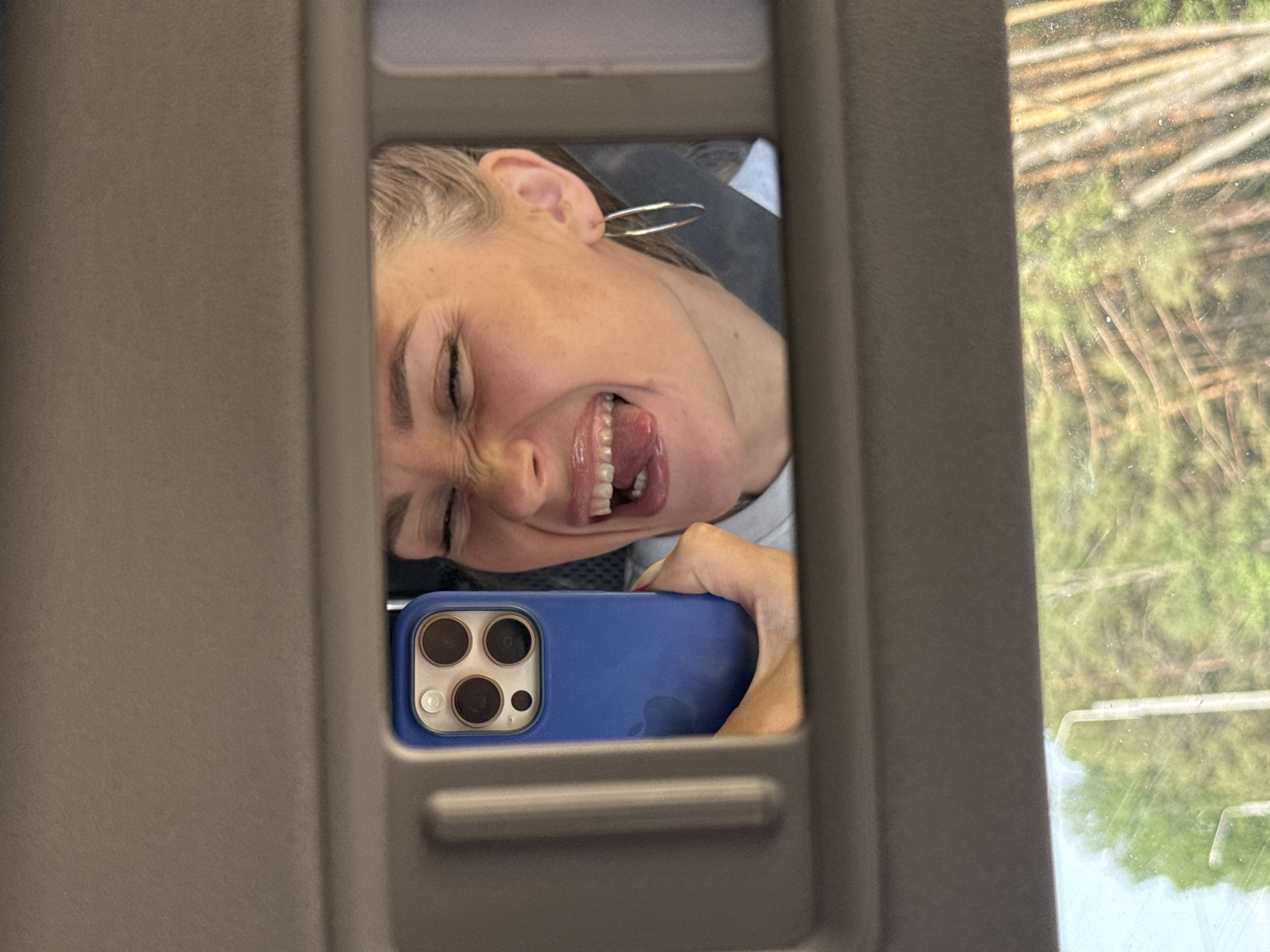 A woman is taking a selfie in the vanity mirror of a vehicle, likely a car, with a blue iPhone. The woman has blonde hair and is sticking her tongue out. A forested area is visible outside the car window. The photo was taken somewhere near the city of Chaikovsky, Russia.