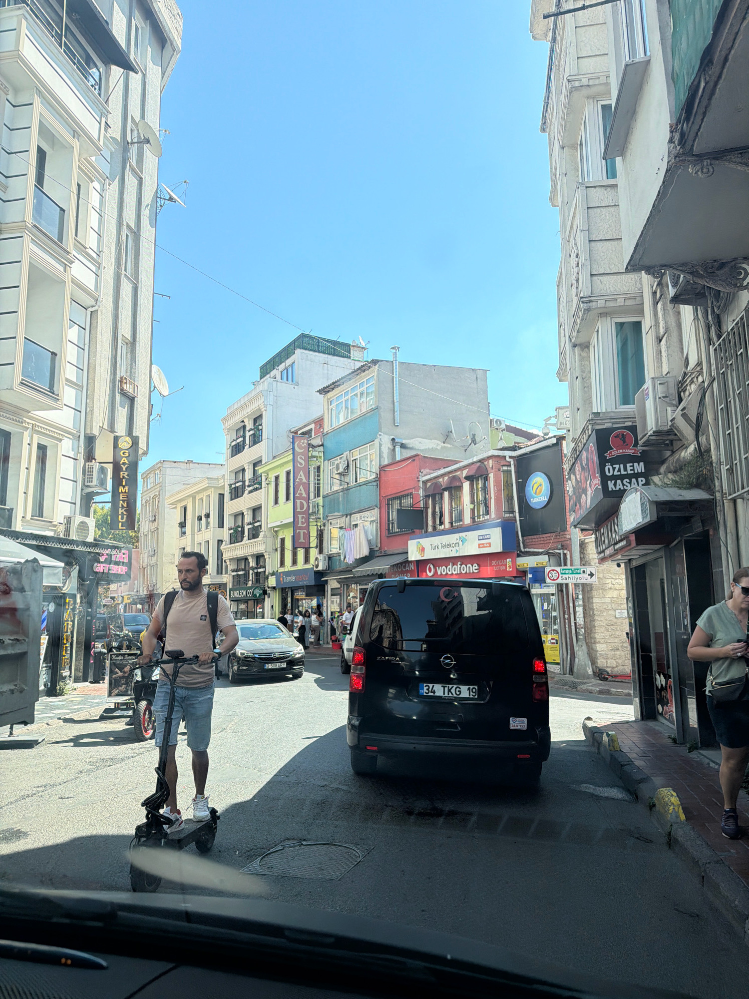 A street scene in Istanbul, Turkey, is captured on a sunny day. The image focuses on a man riding an electric scooter in the foreground, wearing a light-colored t-shirt and denim shorts. He is looking towards the right of the frame. Behind him, traffic is present, including a black minivan with a Turkish license plate reading "34 TKG 19". Other vehicles and pedestrians are visible further down the street.
The buildings lining the street are typical of Istanbul, with varying architectural styles, colors, and heights. Signs for businesses such as "Vodafone", "Türk Telekom", and "Özlem Kasap" are displayed. The street appears to be a commercial area, possibly with shops and restaurants. The sky is clear and blue, indicating good weather. The perspective suggests the photo was taken from inside another vehicle, looking out.
