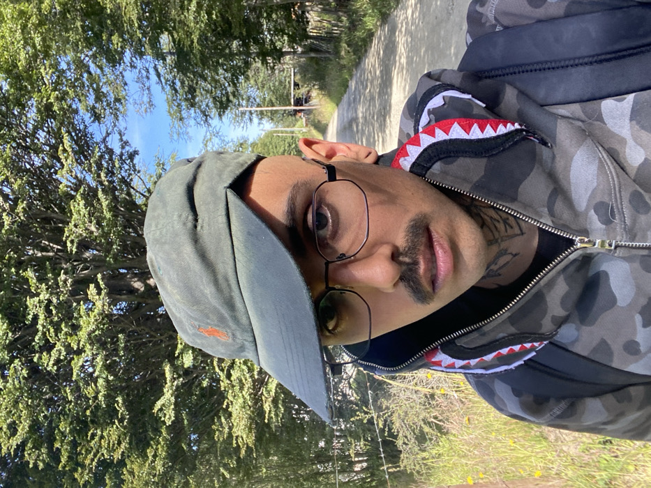 A person wears glasses, a gray baseball cap, and a camouflage shark-motif hoodie, posing outdoors. Trees and a dirt path are visible in the background. The person has a mustache and tattoos on their neck. The photo appears to be taken with a slightly tilted perspective.