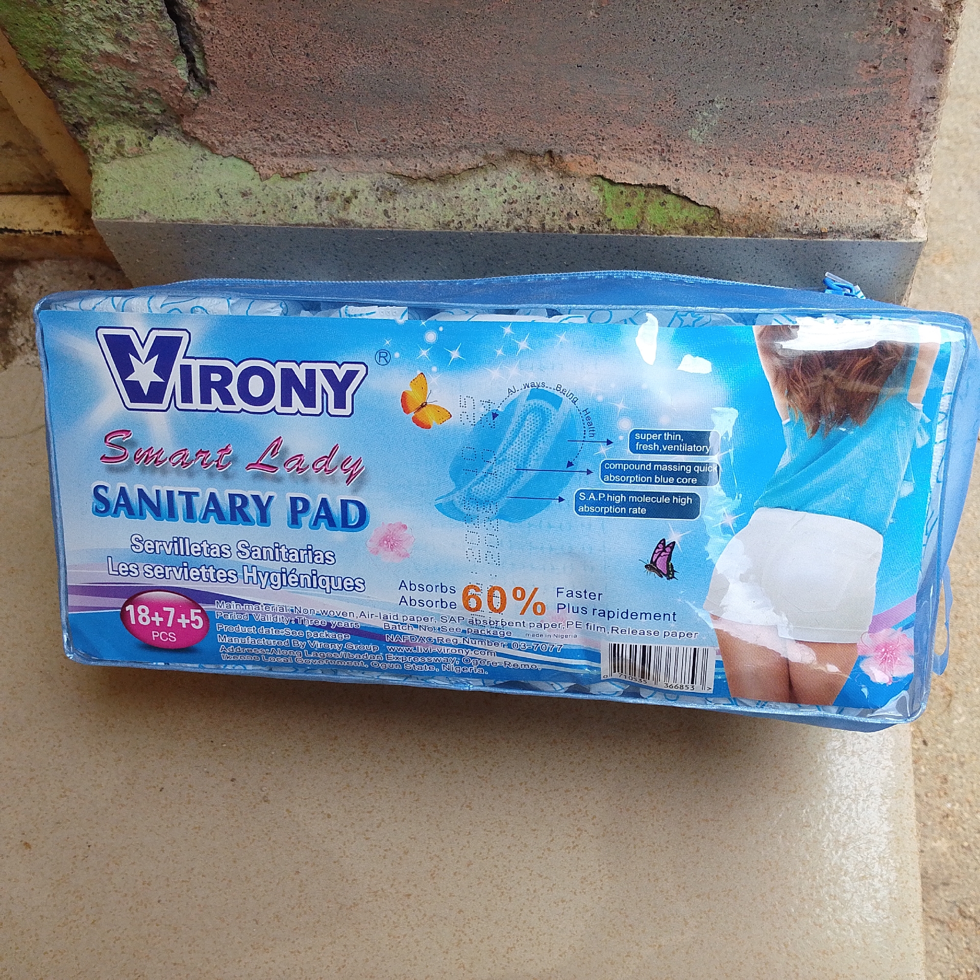 This media file features a close-up shot of a package of "VIRONY Smart Lady SANITARY PADs." The packaging is predominantly blue with white and pink accents, featuring an illustration of a woman from the waist down wearing white underwear. The product name and various descriptive text are clearly visible in English, Spanish, and French. Text highlights include "super thin, fresh, ventilatory," "compound massing quick absorption blue core," and "S.A.P. high molecule high absorption rate." A prominent "60%" symbol indicates the absorption capacity. The package also lists manufacturing details, including the manufacturer "Virony Group," website "www.livi-virony.com," and address in Nigeria. The item is placed on a textured surface, possibly a wall or countertop, with a blurred background that suggests an outdoor or rustic setting with hints of green and brown textures. The lighting appears natural, suggesting it might be daytime. No specific activity or event is depicted beyond the presentation of the product itself.