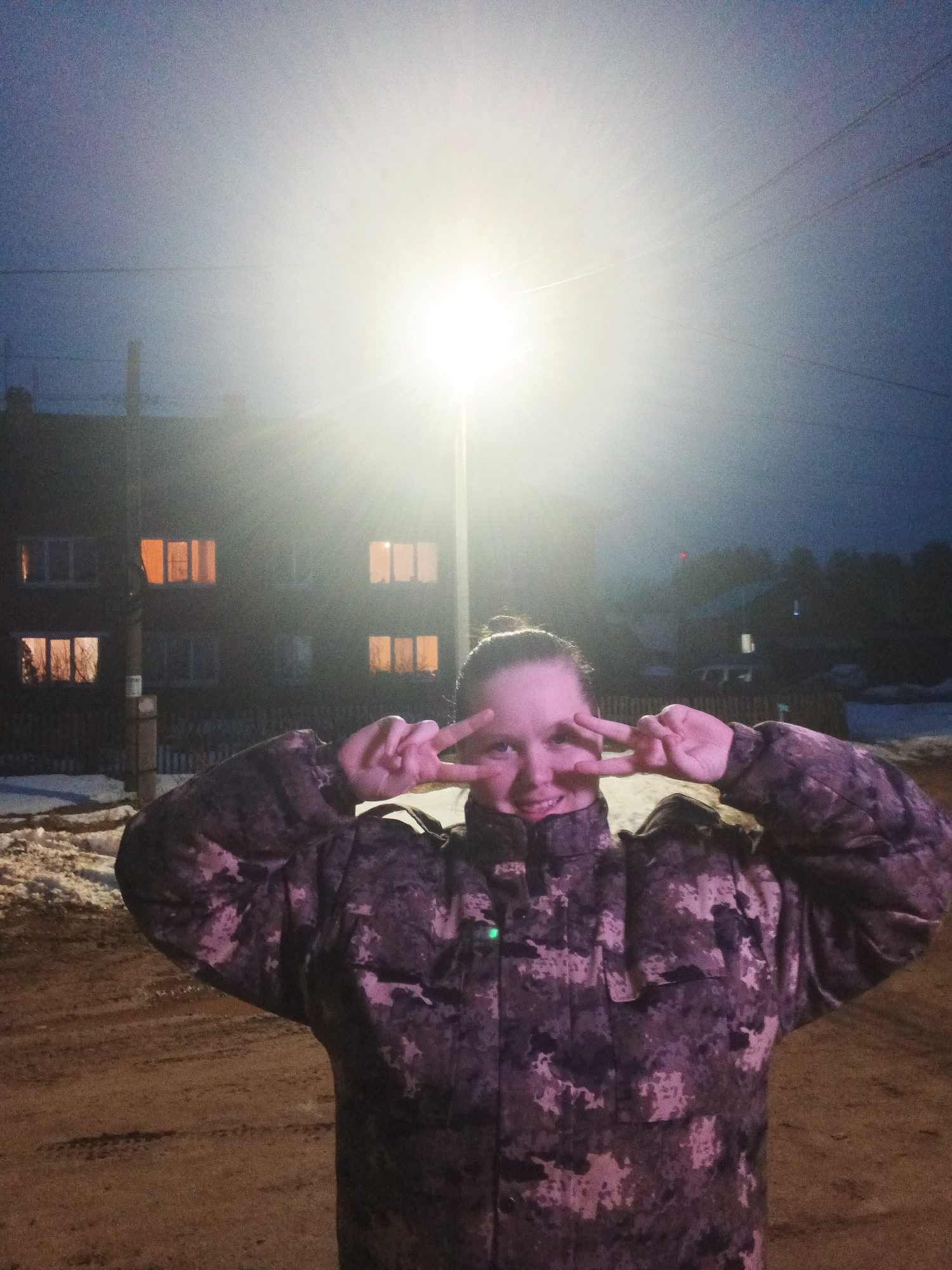 A person is standing on what appears to be a dirt road or path at night. The person is wearing a camouflage-patterned jacket and is making a peace sign with both hands in front of their face. Above the person is a bright street light, which illuminates the immediate area. In the background, there is a two-story building with several windows lit from within. The ground shows patches of snow. Other houses and trees can be discerned in the distance. The image appears to have been taken in Rusalevka, Russia.