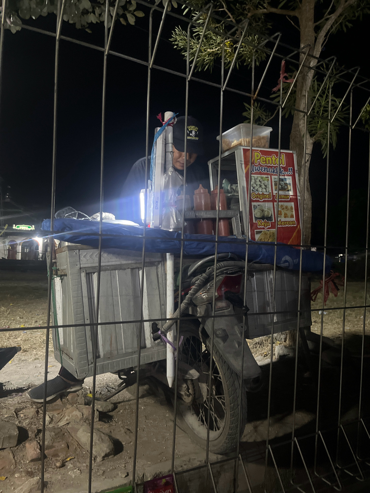 A person is seen behind a metal fence in Banaran, Indonesia, operating a food cart at night. The food cart is attached to a motorcycle and is illuminated by a bright LED light. The cart displays a sign with images of food items and text that appears to be in Indonesian, possibly listing various types of "Pentol" (a type of Indonesian meatball) such as "Original Jamur," "Keju," and "Mercon." Several condiment bottles are visible on a shelf. The person is wearing a dark t-shirt and a baseball cap. The surrounding environment appears to be an outdoor area with some trees and a building visible in the background, dimly lit. The ground is covered with dirt and rocks.