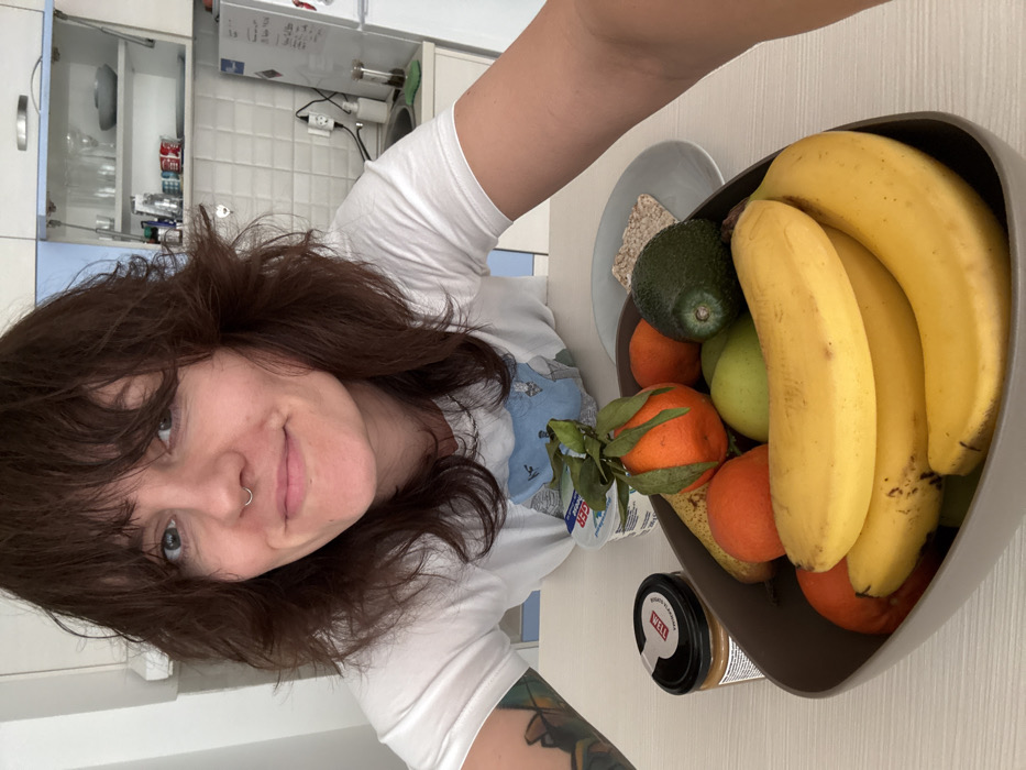 A woman is depicted in a kitchen setting in Boreti, Montenegro, capturing a selfie-style image. The woman has medium-length dark brown hair with bangs, green eyes, and a septum piercing. She wears a white short-sleeved t-shirt with a graphic design. A multicolored tattoo is visible on her left forearm. She is smiling slightly, looking directly at the camera.
In the immediate foreground, on a light-colored countertop, a dark brown, oblong fruit bowl contains multiple bananas, tangerines (one with leaves), green apples, a pear, and an avocado. To the left of the fruit bowl is an open jar of peanut butter with a white and red label (VELL brand visible). Behind the fruit bowl, a white plastic container of yogurt (branded *MEGGI* or similar) is present. To the right, on a small grey plate, are two rectangular crackers.
The background reveals a modern kitchen. White wall cabinets with light blue undersides are visible, one open, revealing glassware and dishes. A white tiled backsplash is present. To the right, a white refrigerator with items attached to its door and a wall-mounted electric socket with a black cord plugged in are visible. A small transparent container with dark contents is on the counter next to the refrigerator. The walls are light-colored.