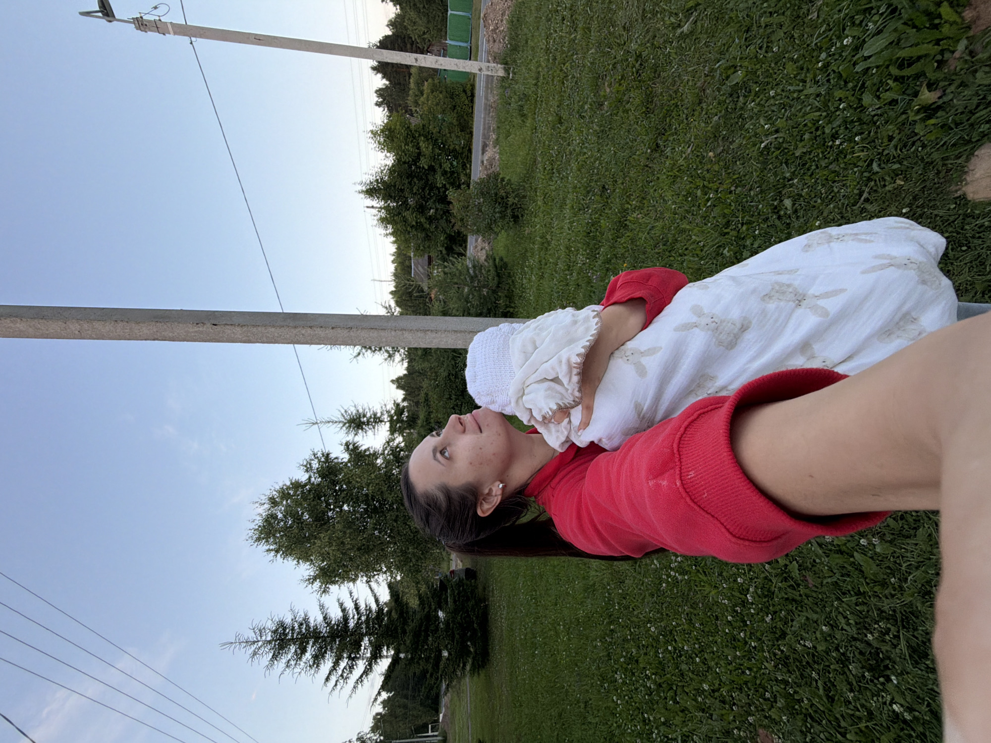 A woman in a red sweatshirt is holding a swaddled baby outdoors. The baby is wrapped in a white blanket with a bunny pattern. The woman is looking up at the sky, and her face is partially visible. They are standing on a grassy area with trees and power lines in the background. A road and a green fence are also visible. The setting appears to be a rural or suburban area.