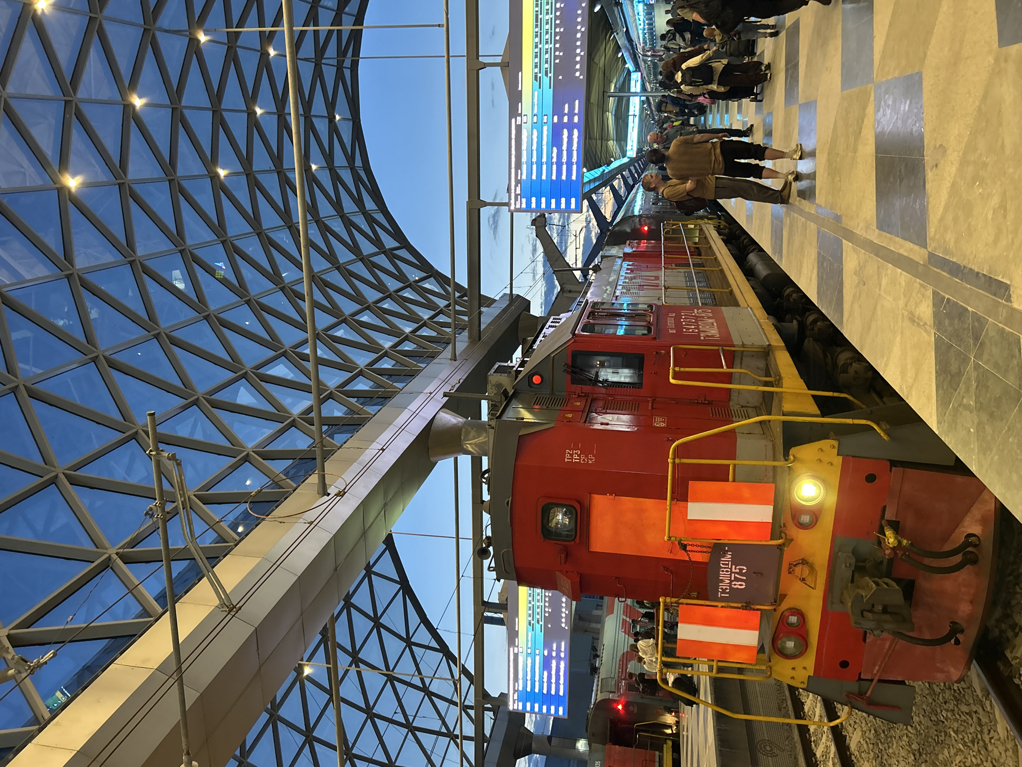 This medium-shot image, taken at a train station, captures a red and yellow locomotive stopped at a platform under a striking modern glass and metal roof structure. The image is oriented with the platform and train running horizontally from left to right.
The most prominent subject is the red locomotive, facing towards the right of the frame. Its front features a bright headlight, yellow handrails, and orange and white striped warning panels. The number "ТЭМ18ДМ-875" is clearly visible in white letters on a red background on its front. The side of the locomotive also displays text, including "ТРАНСЛОКОМ", "ТЭМ18ДМ", and "15473721".
To the right of the locomotive, a tiled platform stretches into the distance, featuring a light and dark checkerboard pattern. Several people are visible on the platform; two men in brown/khaki shirts and dark trousers stand prominently in the mid-ground, appearing to be observing the scene or waiting. Further down the platform, more passengers are gathered, some appearing to be boarding the train or waiting for departure.
Above the platform and train, a large digital information screen glows brightly, displaying what appears to be train schedules or destination information in various colored segments, predominantly blue and yellow. Overhead electrical lines and their support structures are also visible.
The station's architecture is a notable feature, with a vast, curved glass and metal grid roof forming an intricate pattern of triangles. Recessed lights within the structure illuminate the glass. The sky, visible through the glass, is a clear blue, suggesting dusk or early evening, a time of day reinforced by the illuminated screen and station lights. The overall atmosphere is that of a busy, modern transportation hub.