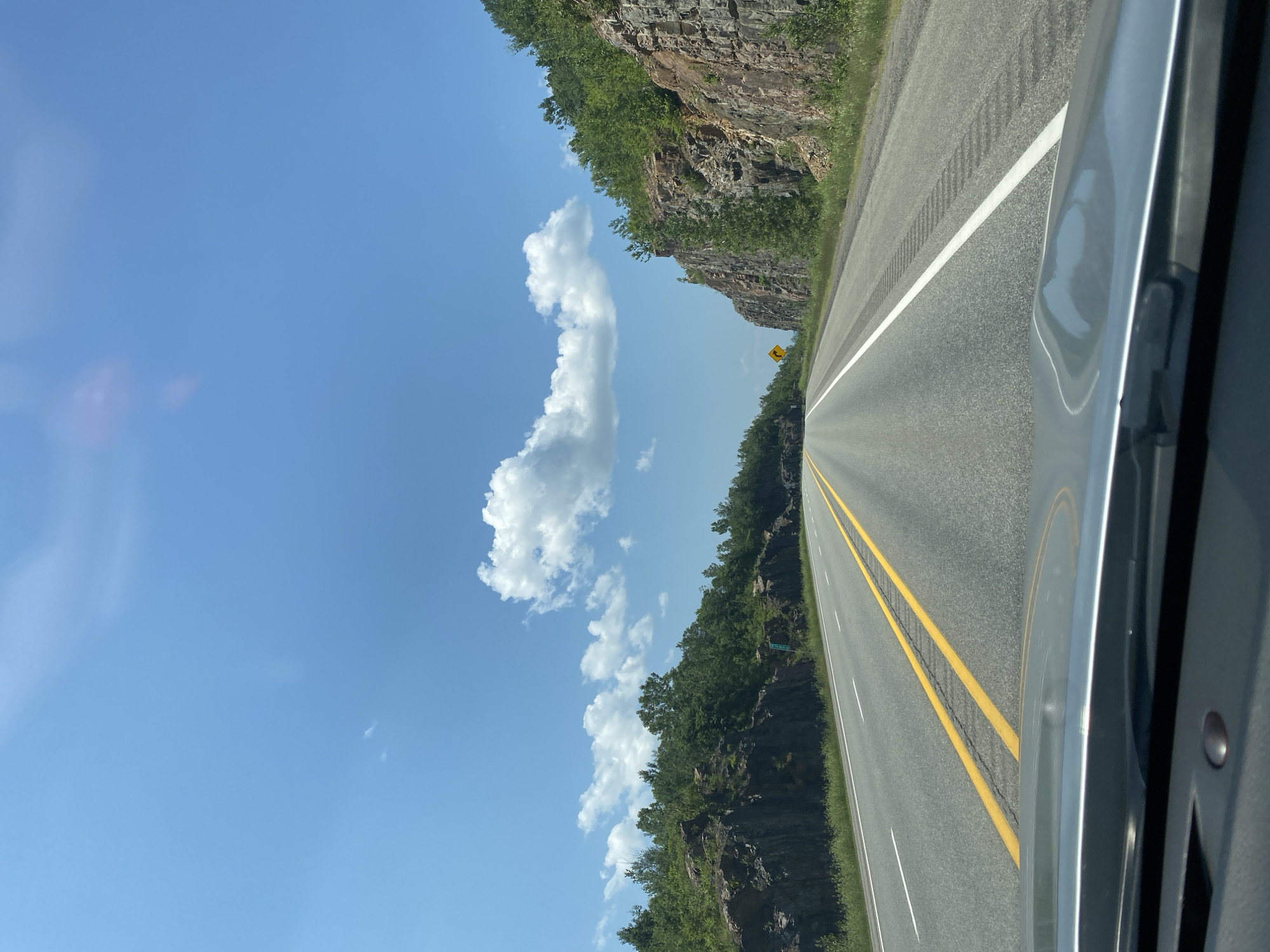 A first-person perspective shot captures a highway road with a bright blue sky and scattered clouds. On either side of the road, there are rocky cliffs with green trees growing on top and along the sides. The road is paved and has yellow and white lines marking the lanes. In the distance, the road curves to the right, disappearing from view. There is a yellow warning sign on the right side of the road in the distance, indicating a curve to the right. The foreground shows the dashboard of a car. There are no people or animals visible in the image. The scene appears to be a natural landscape with a man-made road.