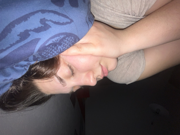 A person lies sideways with their head under a blue patterned blanket. Their eyes are closed, and their face is turned towards the viewer. They have brown hair, fair skin, and a neutral expression. Their arm is bent, with their hand near their neck. They wear a gray top. The background is a mix of light wall and dark space.