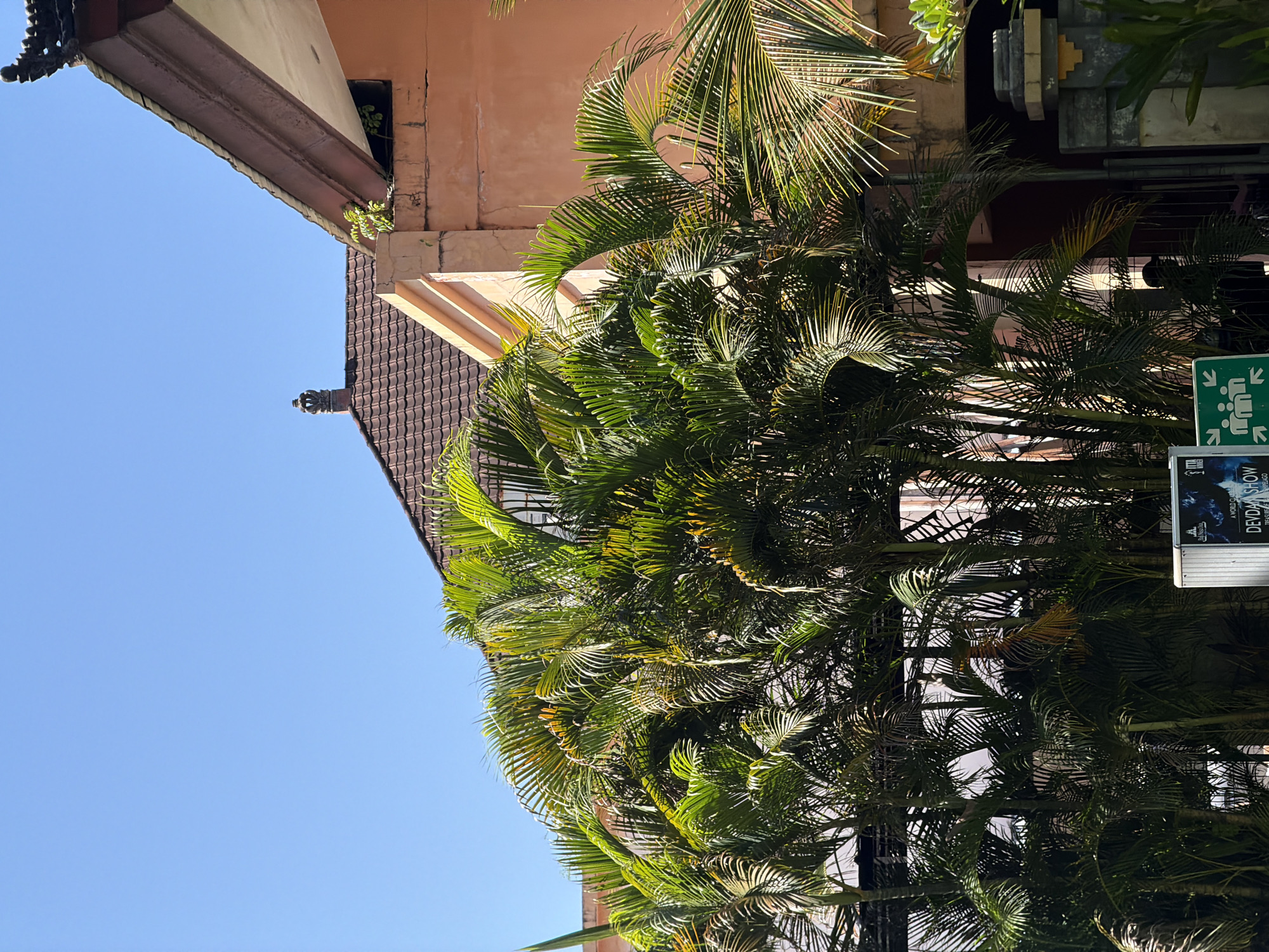 The image shows a view of a building with traditional Balinese architecture, featuring tiled roofs and ornate roof edges. Lush palm trees with vibrant green fronds dominate the foreground, obscuring parts of the building. A clear blue sky forms the background. In the lower right corner, a glimpse of a stone gate structure is visible. A sign with white and green graphics is partially visible in the lower center, appearing to indicate a meeting point or emergency exit. The building's exterior walls are painted a light peach color. This scene is located in Kuta Selatan, Indonesia.
