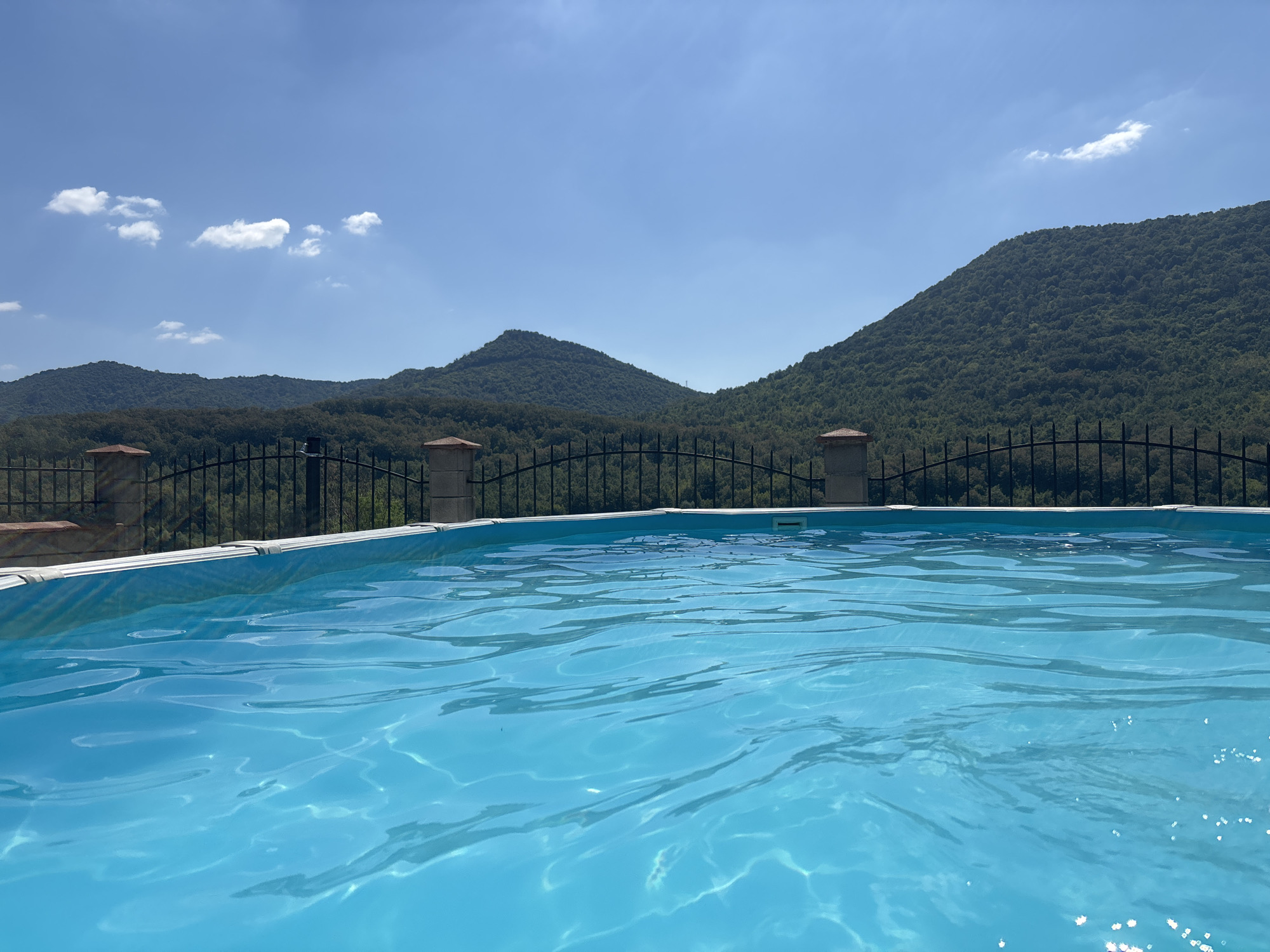 The image shows a swimming pool with bright blue water in the foreground, reflecting sunlight and creating ripples on the surface. The pool is bordered by a white edge. Beyond the pool, there is a black metal fence with decorative posts topped with reddish-brown caps. In the background, rolling green hills covered in dense forest rise towards a clear blue sky with a few scattered white clouds. The scene appears to be a serene outdoor setting. The location is identified as Asparuhovo, Bulgaria. There are no people or animals visible in the image.