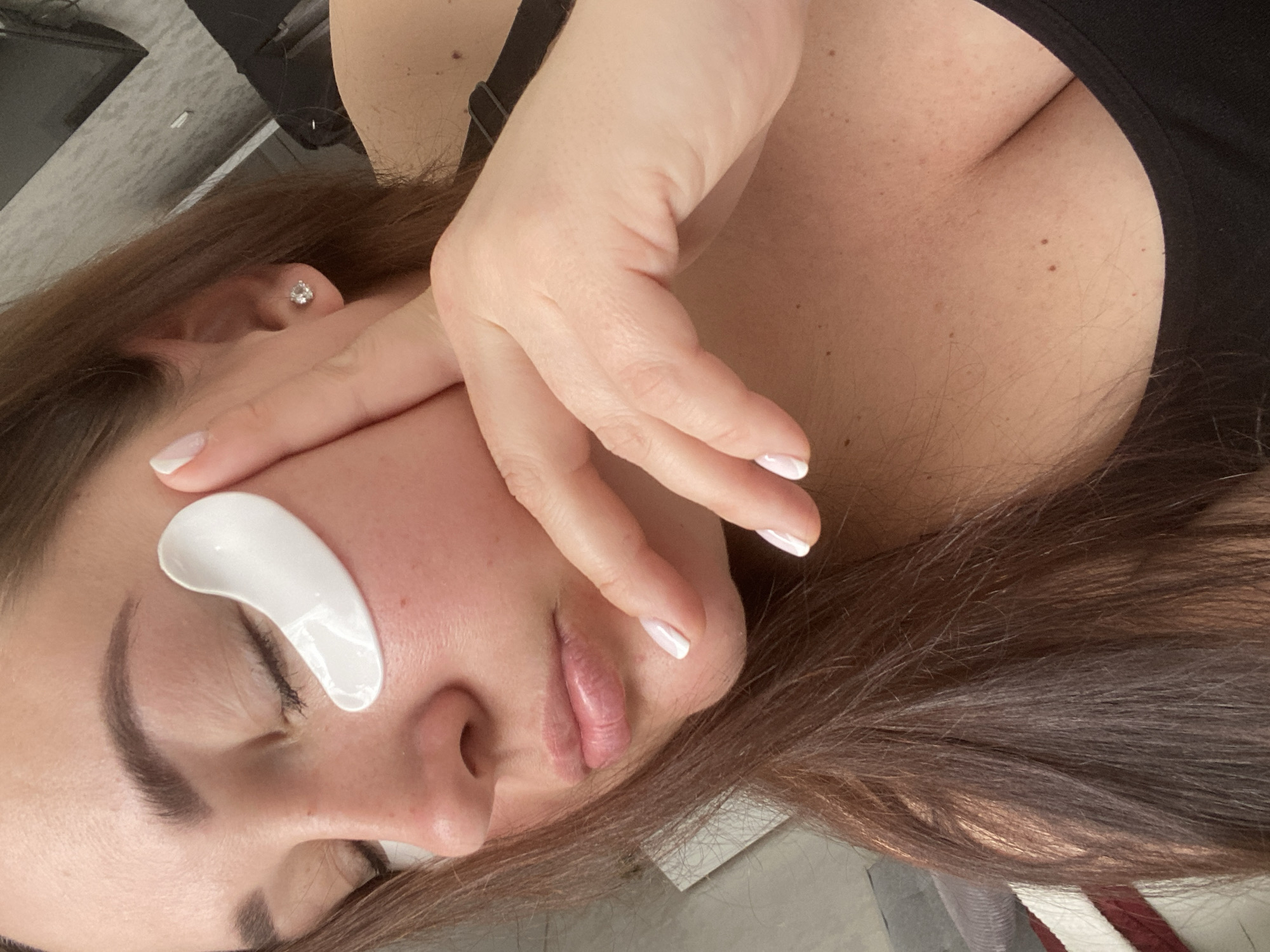 A woman is lying down, eyes closed, with a white eye patch under her left eye. Her right hand is touching her face near her lips. She has brown hair and a light complexion with moles on her chest. She is wearing a black top. Her fingernails are painted white. The image is taken from an overhead perspective. The location is unknown and there is no information available to determine the city of Sochi, Russia.