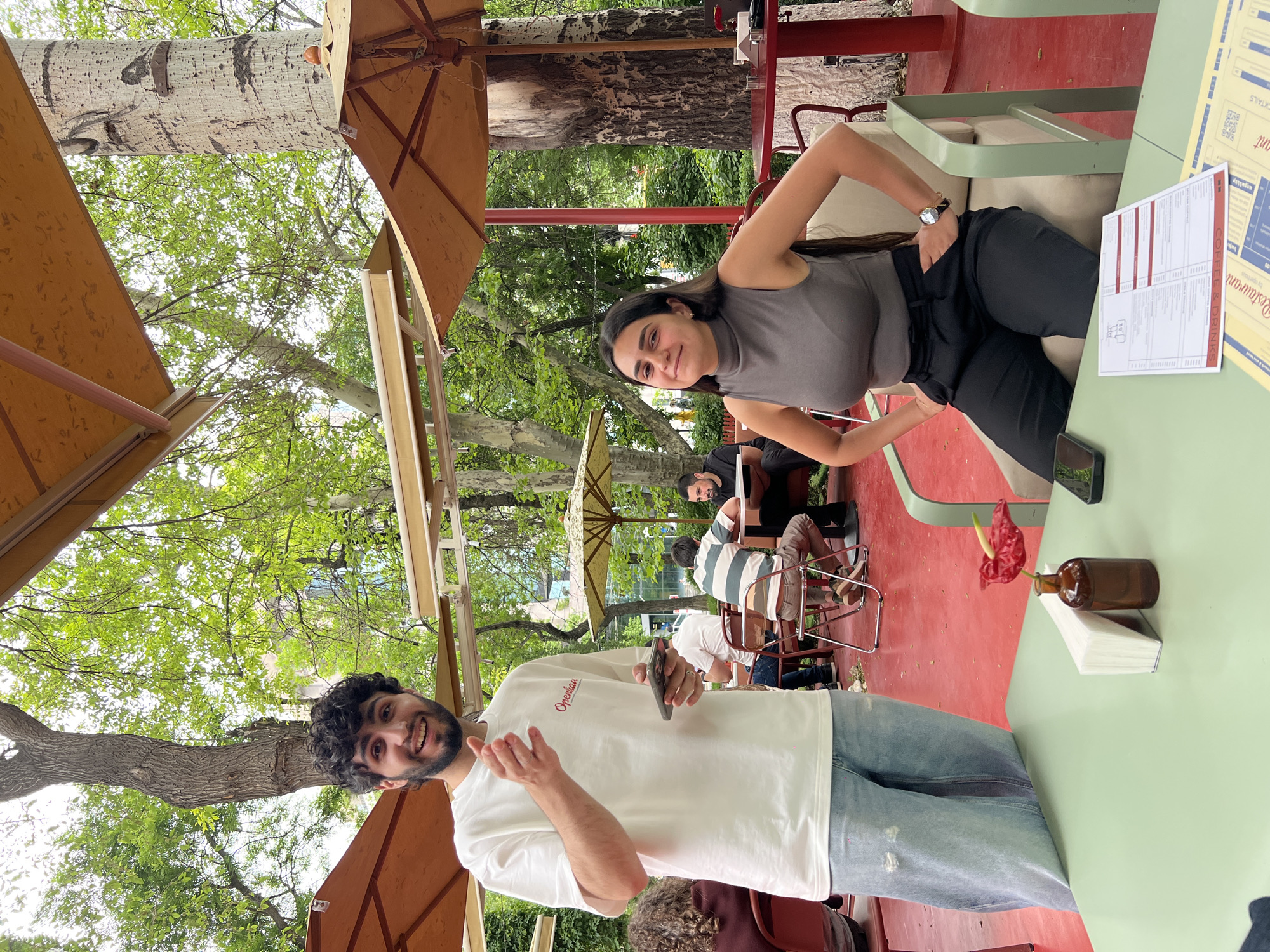 The image captures a scene in an outdoor cafe, likely in Yerevan, Armenia. A man with dark curly hair and a beard, wearing a white t-shirt and light blue jeans, stands at the lower left corner of the frame, smiling and gesturing with his hands. A woman with long dark hair, wearing a gray tank top and black pants, is seated in the background, also smiling. They appear to be posing for a photo. The cafe features green tables and red flooring. A small brown vase with a red flower and a stack of white napkins is placed on the table near the man. In the background, other patrons are seated at tables under umbrellas. The setting is shaded with trees and umbrellas, indicating an outdoor location. A restaurant menu is on the table.