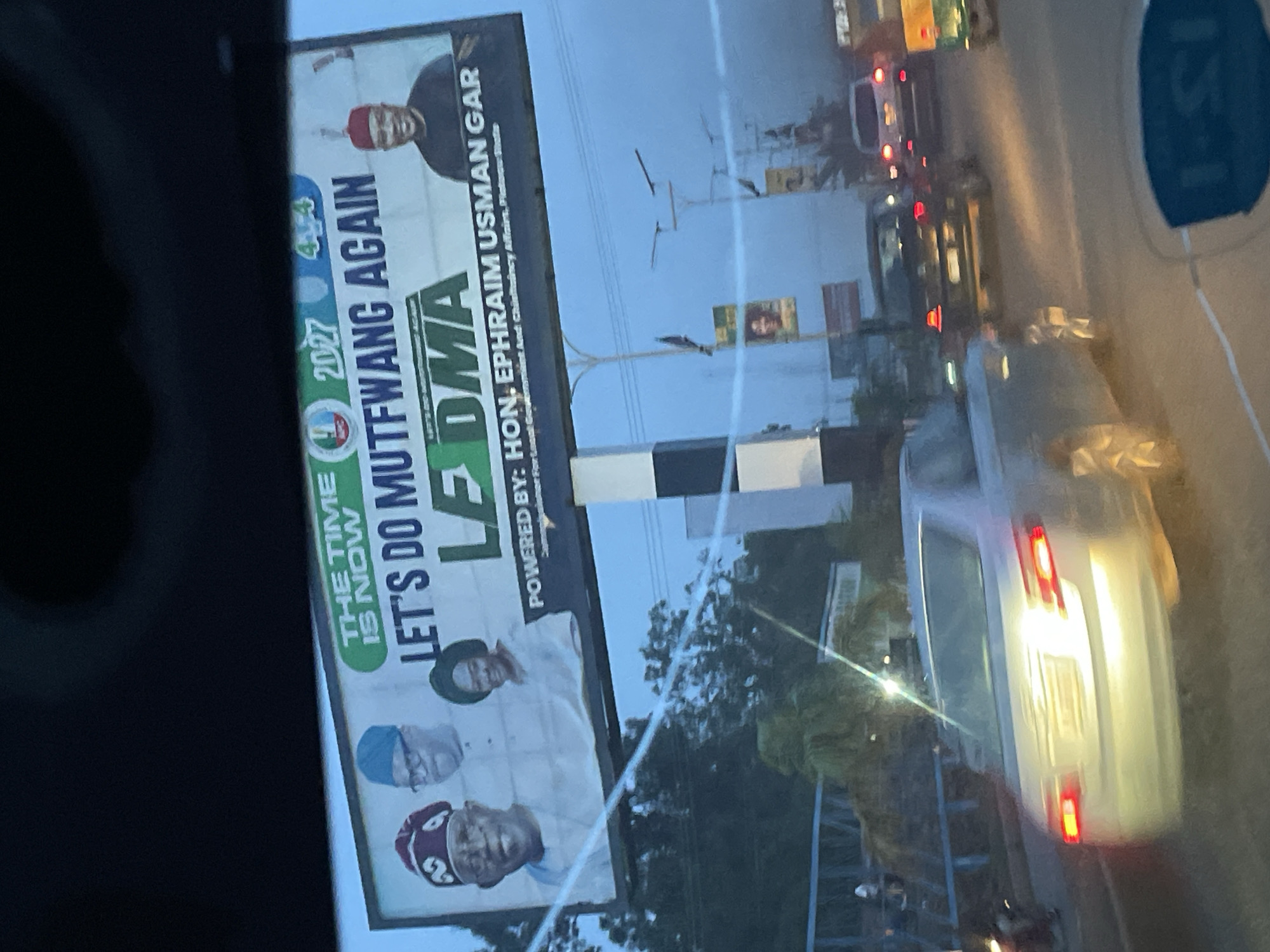 The image is a photograph taken from inside a vehicle, looking out at a billboard in Jos, Nigeria. The billboard displays portraits of three individuals: a man in a red cap and white attire on the left, a man in a blue cap and glasses in the center, and a woman in a green headscarf on the right. The text on the billboard reads "THE TIME IS NOW LET'S DO MUTFWANG AGAIN". Below this, "L SMA" is prominently displayed, followed by "POWERED BY: HON. EPHRAIM USMAN GAR Commissioner For Legal, Government And Chieftaincy Affairs, Plateau State."
The billboard is situated above a street with traffic. Several vehicles, including cars and a motorcycle, are visible on the road. The sky is overcast, suggesting it might be early morning or late afternoon. The overall scene captures a moment of urban life in Jos, with a political campaign advertisement as the focal point.