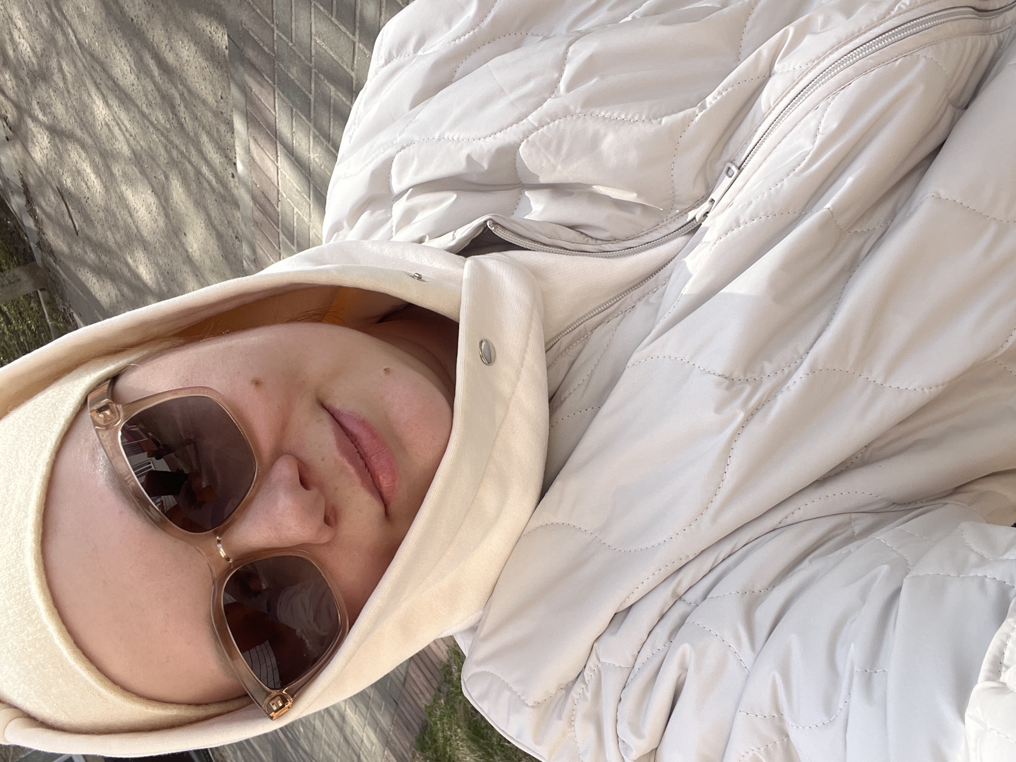 A woman with fair skin, wearing sunglasses and a white hooded jacket, is depicted in an overhead selfie. The background shows a sidewalk, suggesting an outdoor location.
There is no information to suggest the location is Usman, Russia.