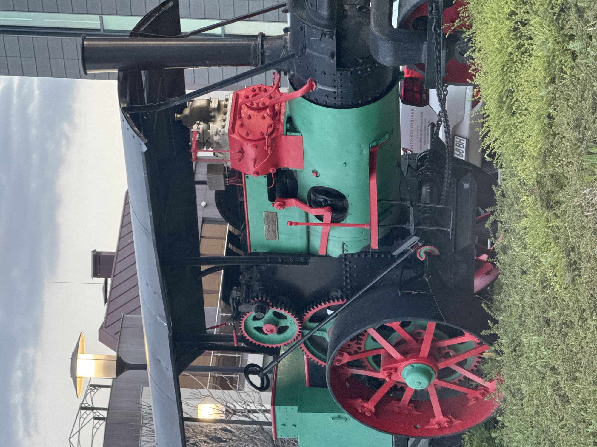 The image depicts a static display of a vintage steam engine, likely a traction engine, located in Varna, Bulgaria. The engine is painted in green, red, and black, with large wheels that have red spokes and green hubs. The engine's body is primarily green, with red components on the top, and black detailing around the smokestack and other mechanical parts. The display is positioned near a modern building with a glass facade and a lamppost, suggesting an urban setting. To the right, a dense bush partially obscures the engine's rear, revealing a license plate number 68 BH. The lighting indicates either late afternoon or early morning, with the sky showing light cloud cover. The scene appears to be a historical exhibit set within the city of Varna.