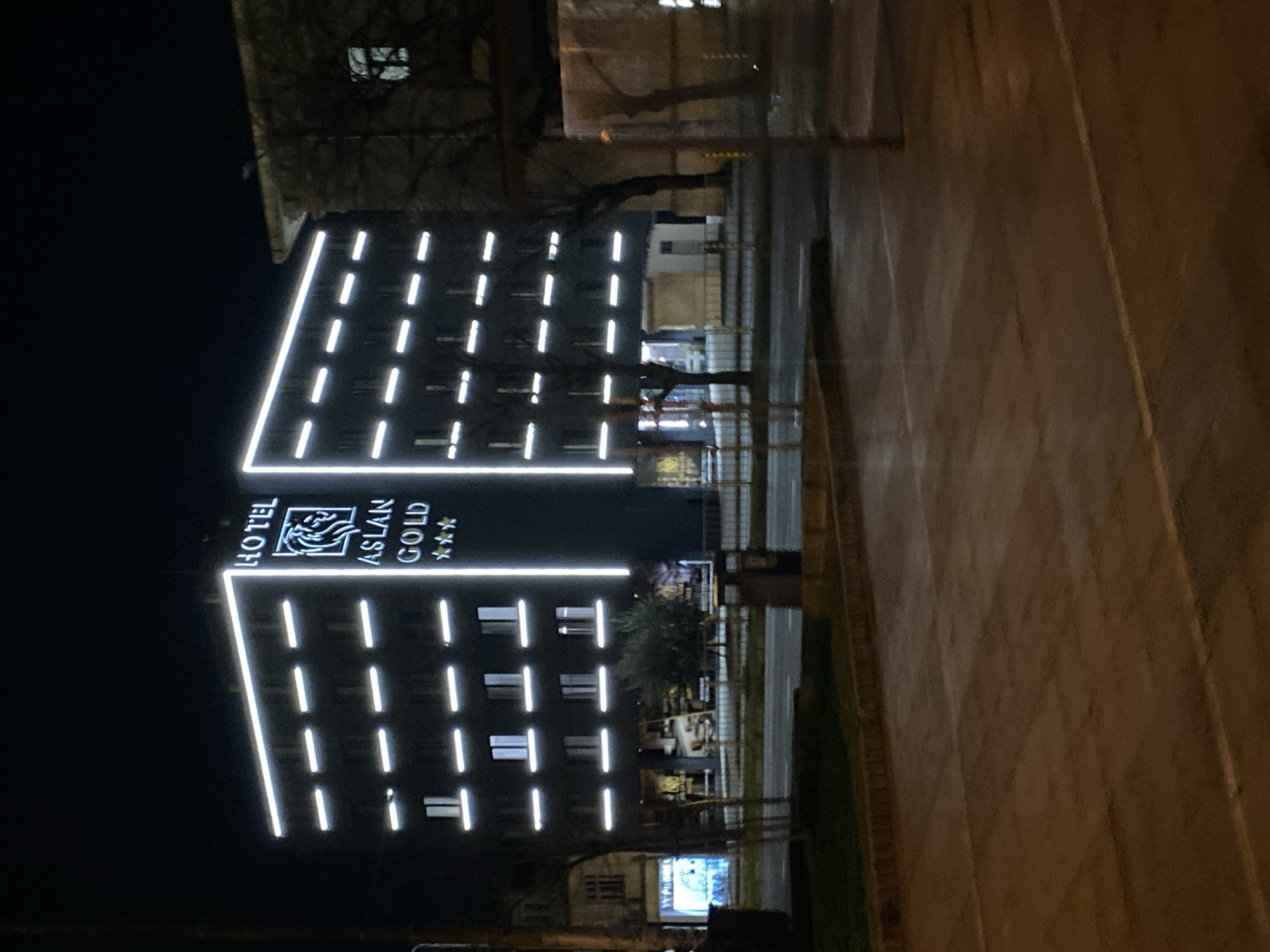 Captured at night in Varna, Bulgaria, the image features the Hotel Aslan Gold, a modern building illuminated with linear white lights. The hotel's name and logo are prominently displayed. The view includes a portion of the street and surrounding buildings. Details include street furniture and hints of tree branches. The overall scene suggests a quiet urban setting at night.
