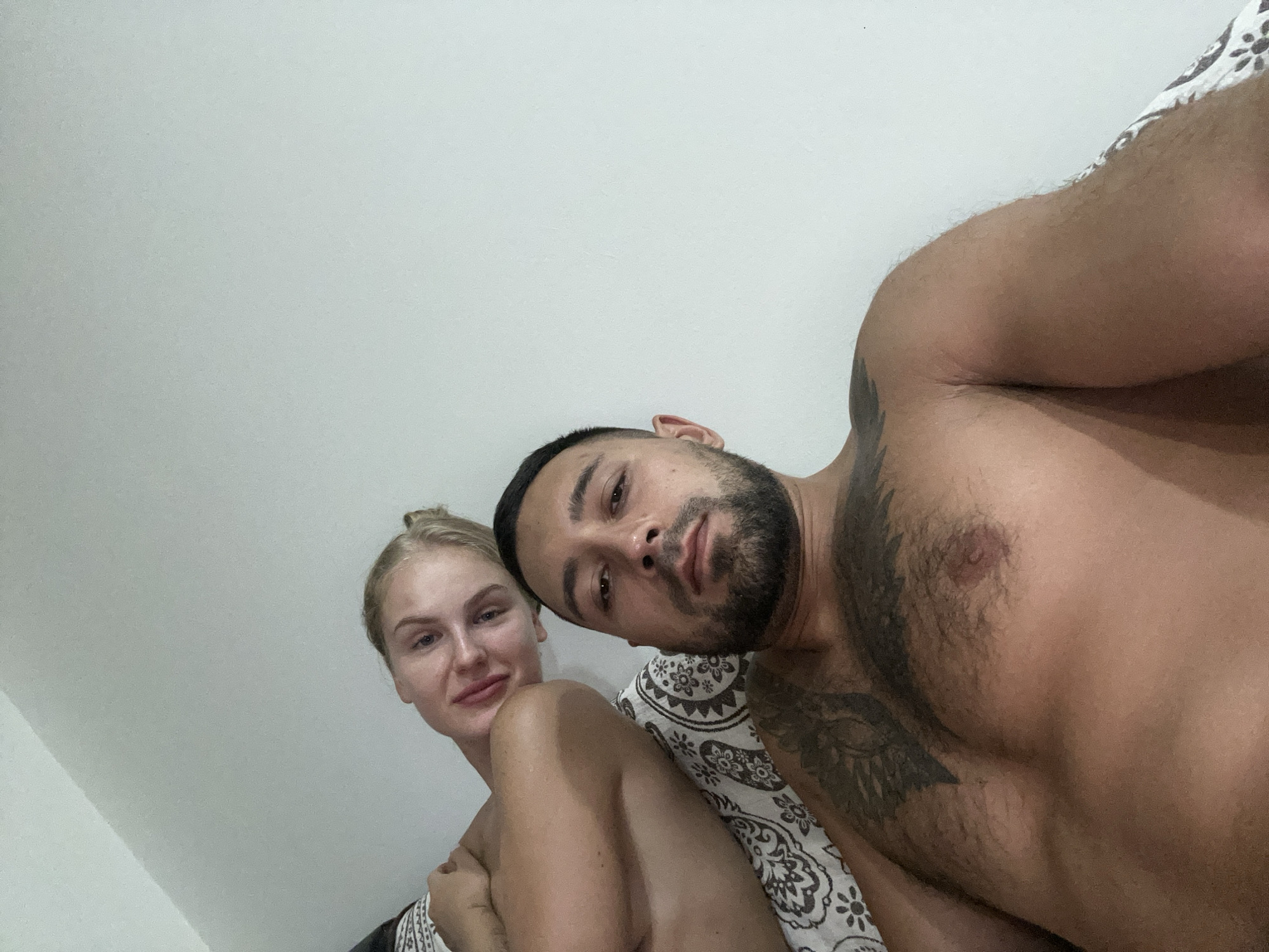 The image shows a close-up, low-angle selfie of a man and a woman. They are lying down, presumably on a bed, as indicated by a patterned fabric visible on the left side of the frame. The man is in the foreground, facing the camera with a beard and chest tattoos. The woman is behind him and to his left, with blonde hair tied up and leaning on him. The background is a plain white wall. The location is specified as Nha Trang Ward, Vietnam.