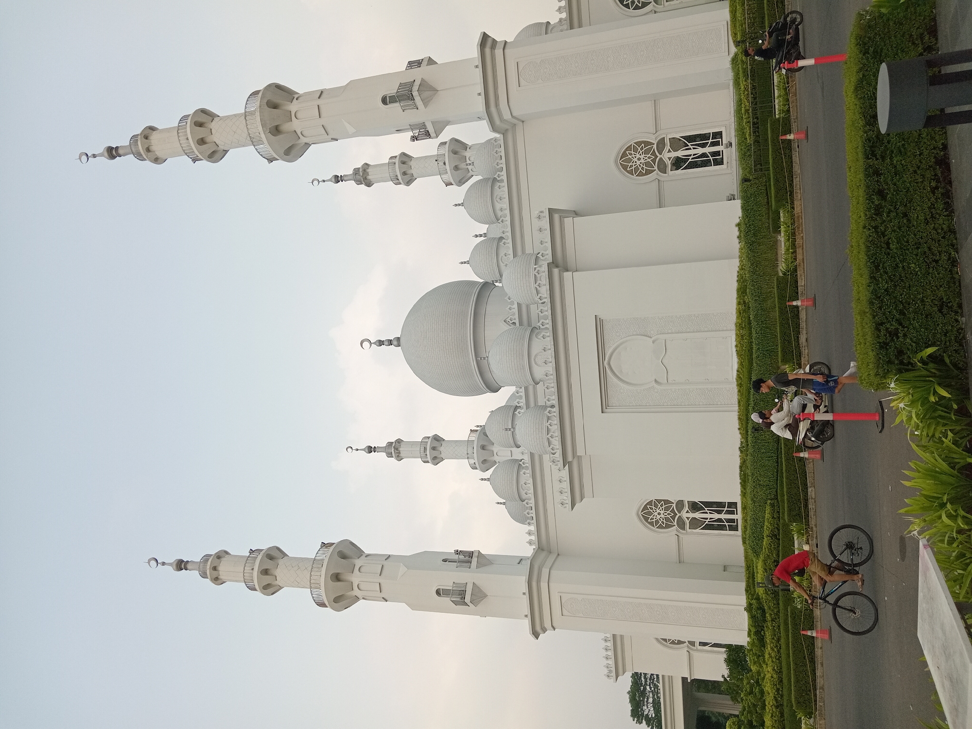 The photograph captures the Grand Mosque of the Indonesian Nawawi Islamic Boarding School in Gunung Putri, Indonesia. The mosque is a large, white structure featuring multiple domes and minarets, with intricate decorative elements. In the foreground, people are seen engaging in various activities: a man is cycling on the left, while two other individuals are riding a motorcycle in the middle of the frame, and another person is on a motorbike on the right. The road in front of the mosque is lined with neatly trimmed hedges and orange traffic cones. The sky above is overcast.