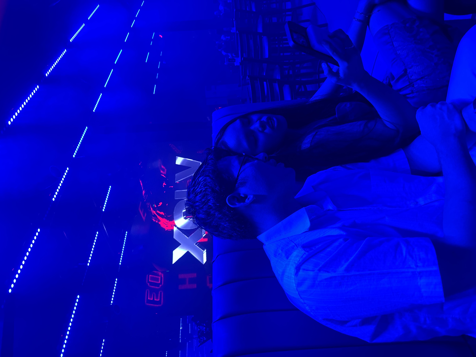 The image shows a dimly lit nightclub scene in Davao City, Philippines, dominated by vibrant blue lighting. Two people are the primary subjects. On the right, a person, possibly male, is lying down and looking at their phone. They are wearing a white collared shirt. Next to them, a person, possibly female, is also lying down, with their head resting on a surface. Their face is partially illuminated, and they appear to be looking in the same direction as the person on the right. Behind them, a large neon sign is visible, with the letters "H OX" illuminated in white and red. Other neon signs with red lettering are also present in the background. The scene is characterized by the intense blue ambient light and the bright neon signs, typical of a nightlife venue. The atmosphere appears relaxed, with the subjects engaged with their phones and seemingly comfortable in the environment. The presence of the "H OX" sign suggests a specific venue or event, though its exact meaning is not clear from the image alone. The overall impression is one of a casual evening out in a lively urban setting.