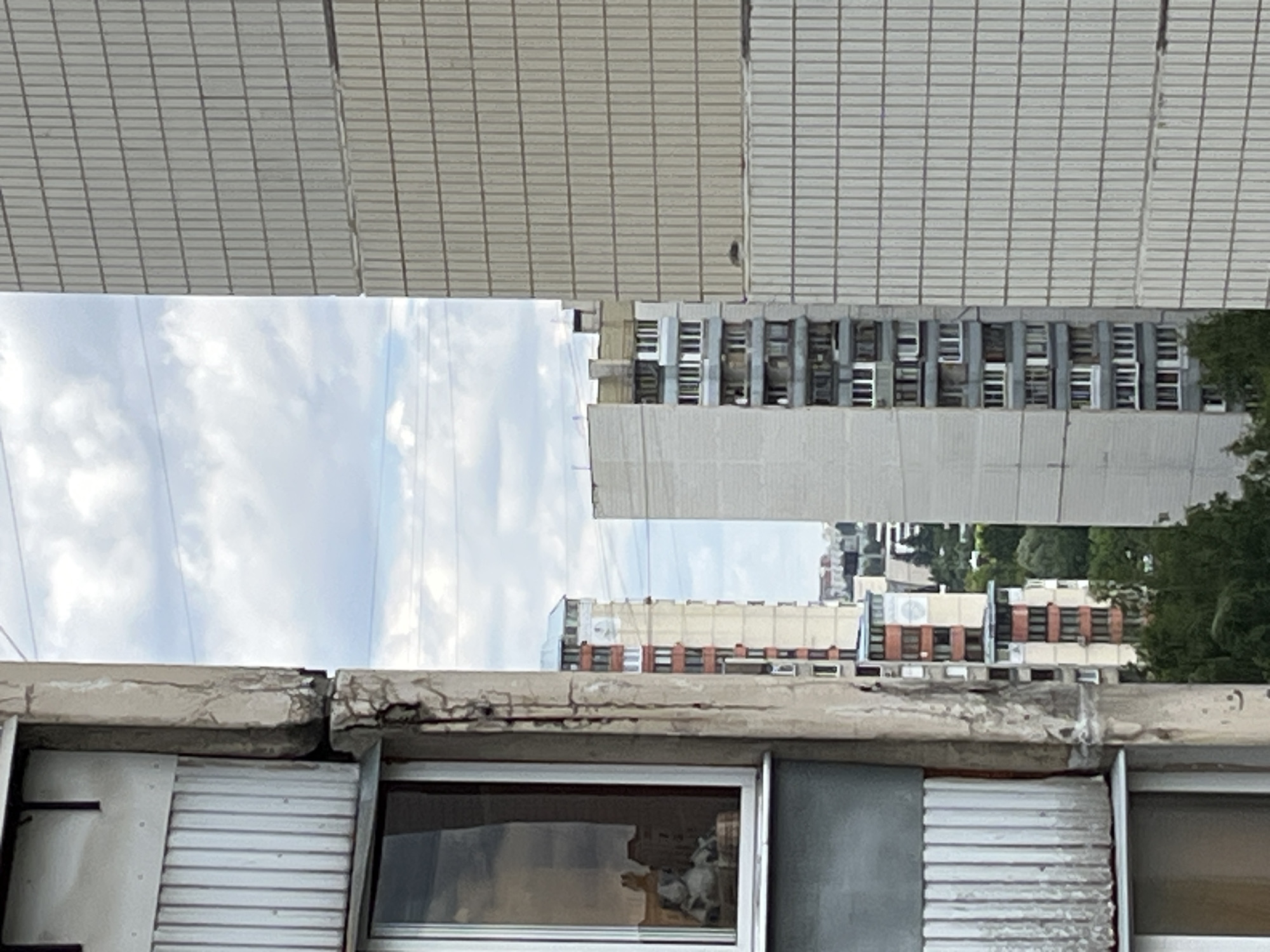 The image shows a view of multi-story residential buildings from a window, with a cloudy sky in the background. The closest building, on the left, has a weathered concrete facade with visible cracks and vertical ribbed panels. A window on this building reflects parts of the interior, including what appears to be decorative objects.
In the middle ground, several other apartment buildings are visible. One is a tall, slender building with a light gray facade and numerous regularly spaced windows, some with darker frames. To its left is another apartment building with a light beige and red-orange color scheme, featuring balconies and distinct sections. Further back, smaller buildings and trees are visible, suggesting an urban residential area.
The building on the right side of the frame is a large, light gray structure with a textured facade composed of repeating vertical elements. The overall scene depicts a common urban landscape, likely in a residential district of Moscow, Russia, characterized by the typical architecture of Soviet-era or post-Soviet era apartment blocks. The time of day appears to be daytime, with overcast sky conditions. No people or specific actions are discernible in the image.