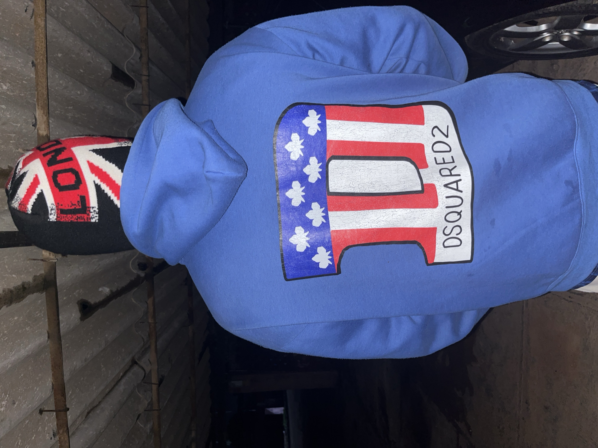 The image shows the back of a person wearing a blue hoodie with a graphic print on it. The graphic is a stylized "D" with red and white stripes, outlined in black, and a blue background with white stars at the top. Below the "D" graphic, the text "DSQUARED2" is printed in black. The person is also wearing a black beanie with the Union Jack pattern and the word "LONDON" in red letters. The background appears to be an outdoor, possibly covered area, with a corrugated metal roof overhead and a dark, possibly paved ground. To the right, a car wheel is partially visible. The image is taken at night or in low light conditions. There is no discernible information in the image that can be used to determine the location as Jos, Nigeria.