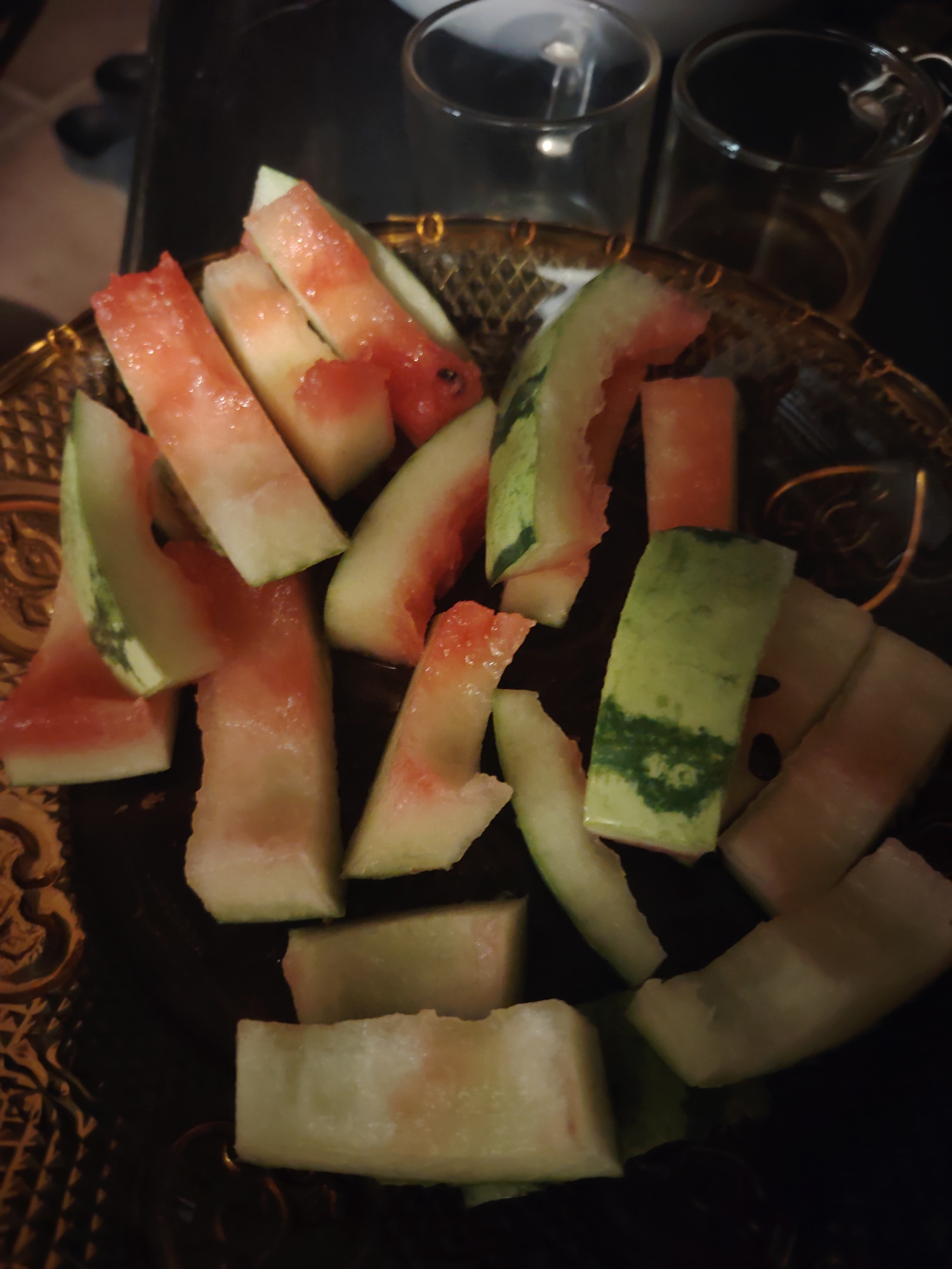 The image displays a close-up, slightly overhead view of numerous discarded watermelon rinds on a dark, ornate plate. The rinds are cut into various irregular strips and block shapes, showcasing the bright green outer skin, the thick white pith, and remnants of pinkish-red watermelon flesh. Some pieces appear glistening with moisture.
The rinds are scattered across a decorative plate that is dark in color, possibly brown or amber, and features an embossed, intricate pattern along its rim and surface. In the blurred background, two empty, clear glass cups or mugs are partially visible, positioned behind the plate to the upper right. The overall background is dark and out of focus, suggesting an indoor setting such as a kitchen counter or dining table, under dim lighting conditions. There is no discernible activity taking place beyond the presentation of the watermelon waste. No people are visible, and there is no visible text. The lighting appears artificial, contributing to the dark and somewhat intimate atmosphere, potentially indicating evening or night.