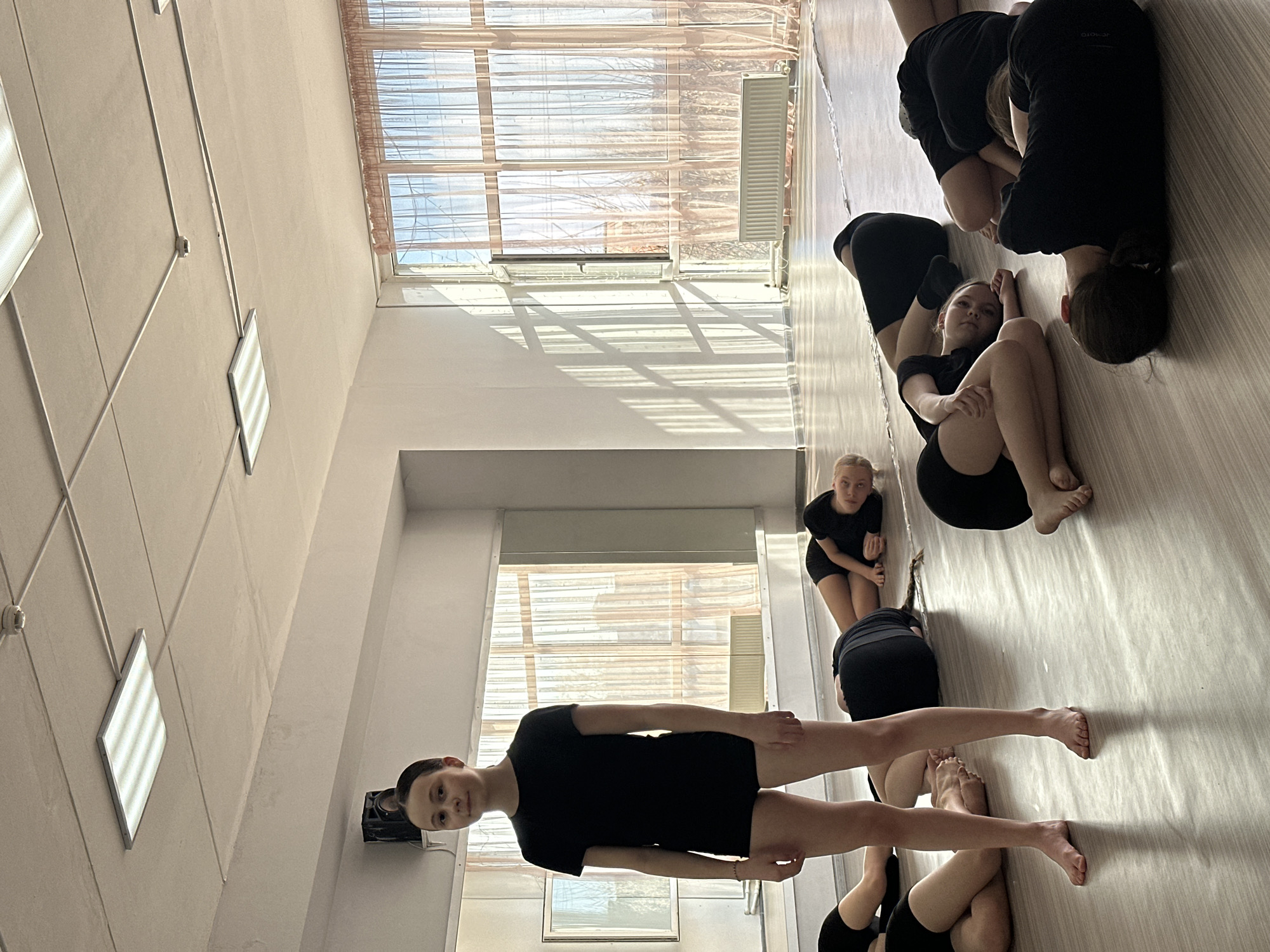 The image depicts a group of young ballet dancers in what appears to be a dance studio in Votkinsk, Russia. The dancers are all wearing black leotards. One dancer is standing straight and looking directly at the camera, while others are sitting on the floor in varying yoga poses, some looking at the camera while some are not.
The dance studio has large windows with sunlight streaming through. The floor is wooden. Fluorescent lights are present on the ceiling. There is a framed picture on the wall behind the standing dancer. A radiator is visible near the windows.