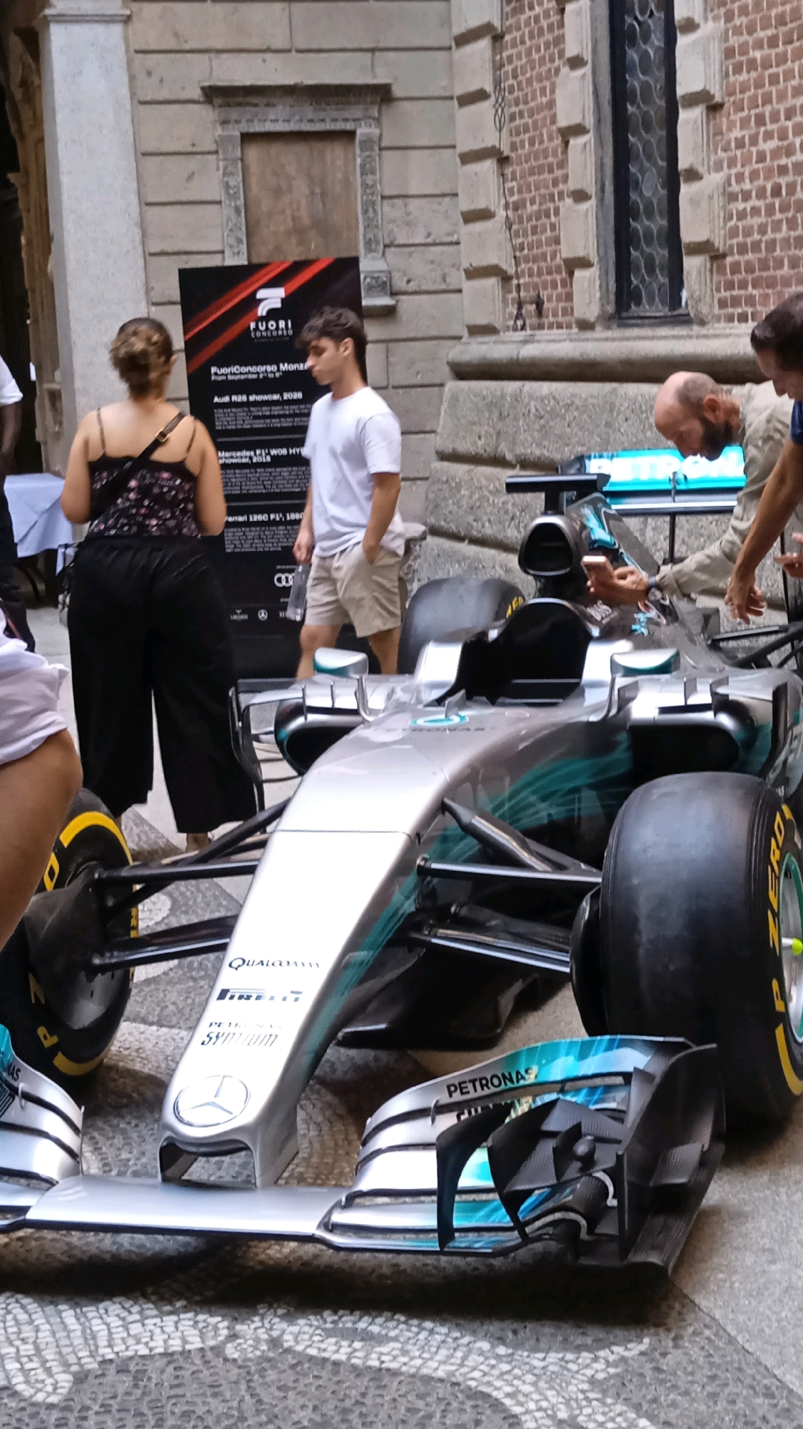 An outdoor street scene in Milan, Italy, features a silver Formula 1 car parked on a patterned stone pavement. The car, bearing the "Petronas" sponsor logo and a Mercedes-Benz emblem, is the central focus. To the left of the car, a woman in black pants and a black patterned top stands with her back to the camera, facing a large black sign. A young man in a white t-shirt and beige shorts stands to the right of the sign, looking towards the right. Further to the right, two men are observing the car, one with a shaved head wearing a beige jacket, and another partially visible man in a blue shirt. Behind the car and to the left, a draped white table is visible. The background consists of a stone building with arched windows, one of which is boarded up. The overall scene appears to be a public display or event.