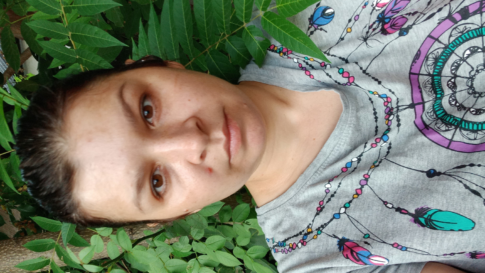 A close-up, waist-up, selfie-style photograph captures a woman with medium-toned skin and dark brown hair pulled back. She is positioned in front of lush green foliage. The woman is wearing a grey t-shirt adorned with a colorful dreamcatcher design, featuring feathers and beads. Her expression is neutral, and her gaze is directed towards the camera. A single pimple is visible on her right cheek, just below her eye. The background consists of dense green leaves and a tree trunk, suggesting an outdoor setting in a park or garden. The lighting indicates it is daytime, with natural light illuminating the subject. The specific location within Kropyvnytskyi, Ukraine, is not discernible from the visual information provided.
