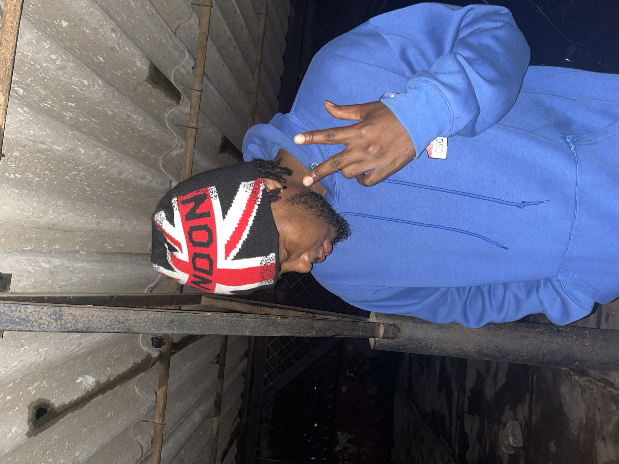 A young Black male wearing a blue hooded sweatshirt and a black beanie with the Union Jack design is standing in front of a corrugated metal roof. He is looking down and to his left, with his right hand raised in front of his chest, displaying a peace sign with his index and middle fingers. The background is dark and appears to be an outdoor or partially enclosed space with visible metal supports and the ribbed texture of the roof. The image is shot from a medium-low angle, focusing on the subject and his immediate surroundings.