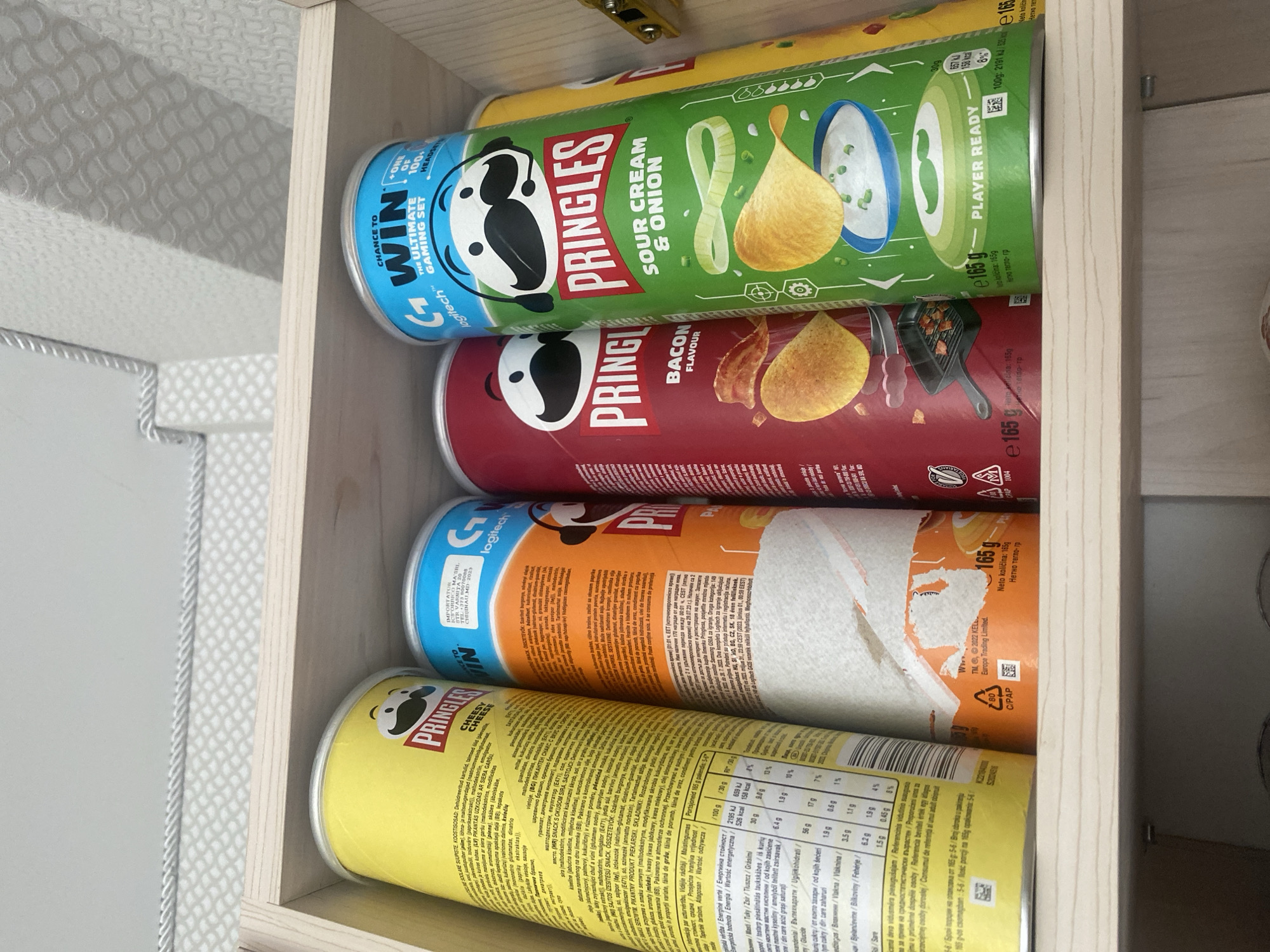 The image shows four Pringles cans lined up on a wooden shelf. From left to right, the cans are: Pringles Cheesy Cheese, Pringles Original, Pringles Bacon Flavour, and Pringles Sour Cream & Onion. The Pringles Original can is a light blue color with a "Logitech" logo, indicating a potential promotion or collaboration. The Pringles Bacon Flavour can is red, and the Pringles Sour Cream & Onion can is green. The wooden shelf is part of a cabinet. The background shows a patterned wallpaper and part of the cabinet door. The lighting suggests an indoor setting, likely a home or storage area. No people are visible in the image. The scene appears to be located in Chișinău, Moldova, based on the presence of the importer's information on the Pringles Original can, which lists "ICS GRINFROD MA' SRL STR. CIUBUK 5/1, MD-2009, CHIŞINĂU, MOLDOVA".