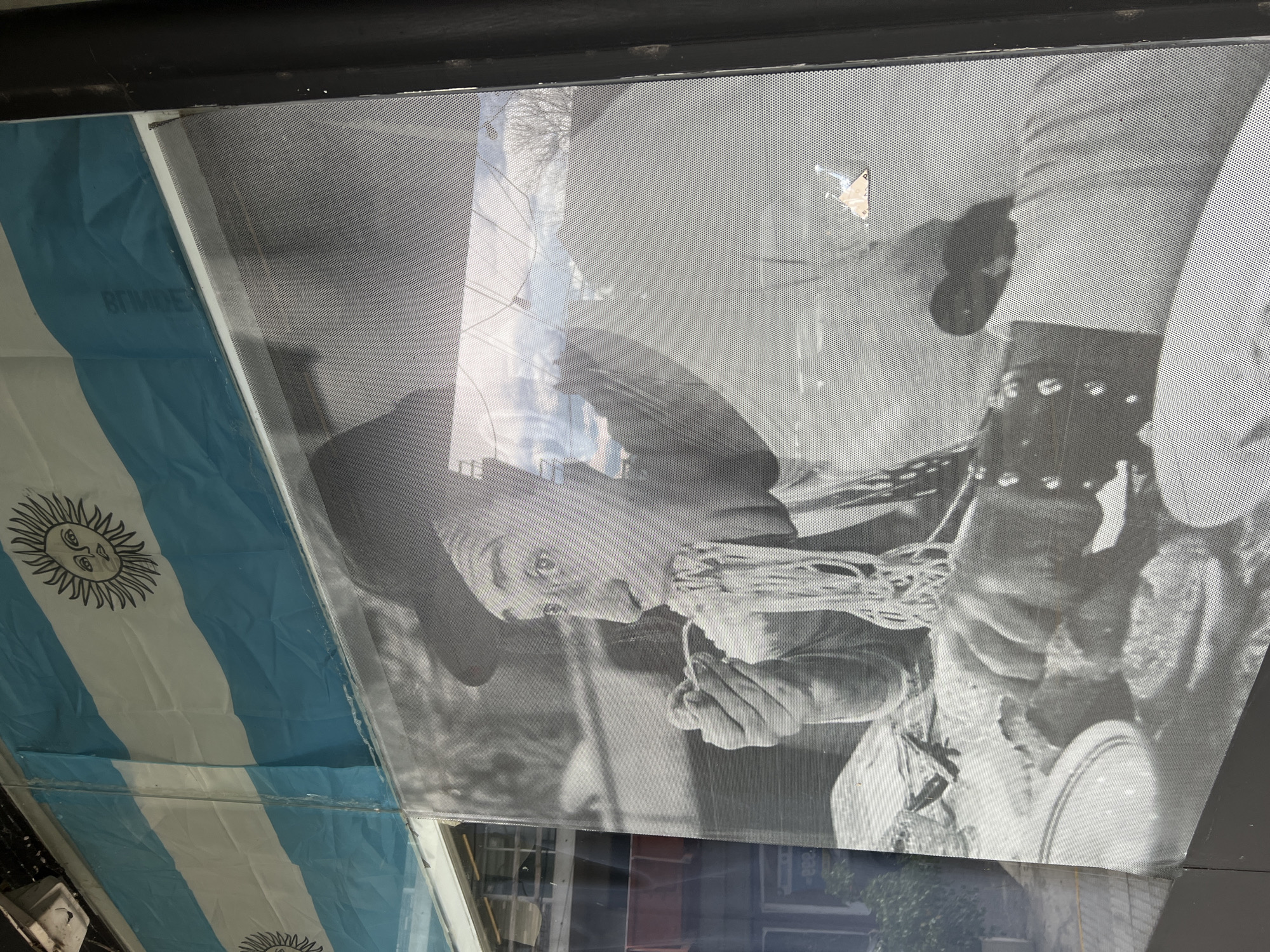 The image shows a shop window in Buenos Aires, Argentina. The window display consists of a perforated film poster featuring a black and white photograph of a man in a cowboy hat eating spaghetti. To the left of the poster, partially obscuring it, is an Argentinian flag. Reflections of buildings and street features are visible on the window.